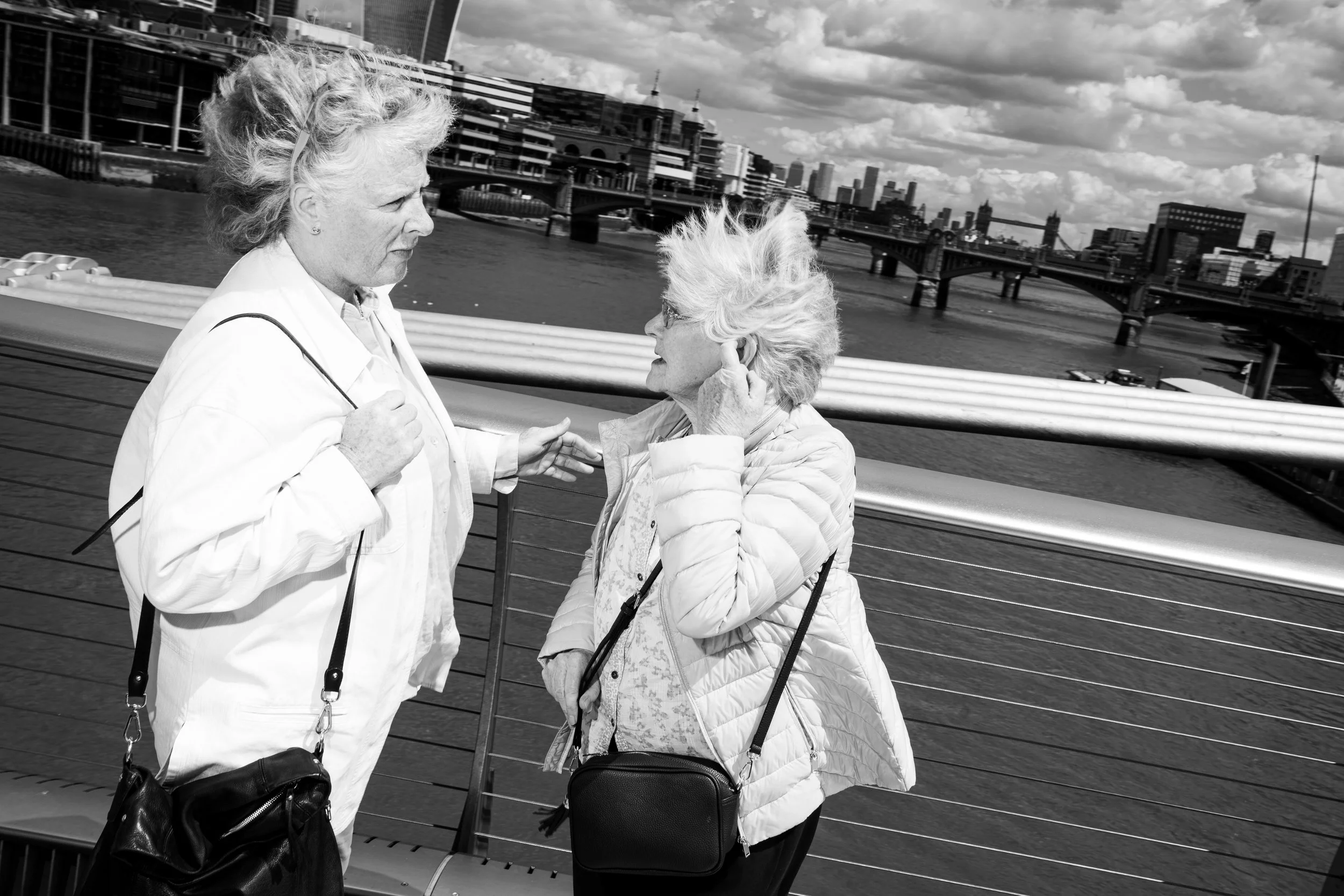 Two elderly women with light-colored hair engaged in conversation on a city bridge over a river, with buildings and bridges visible in the background.