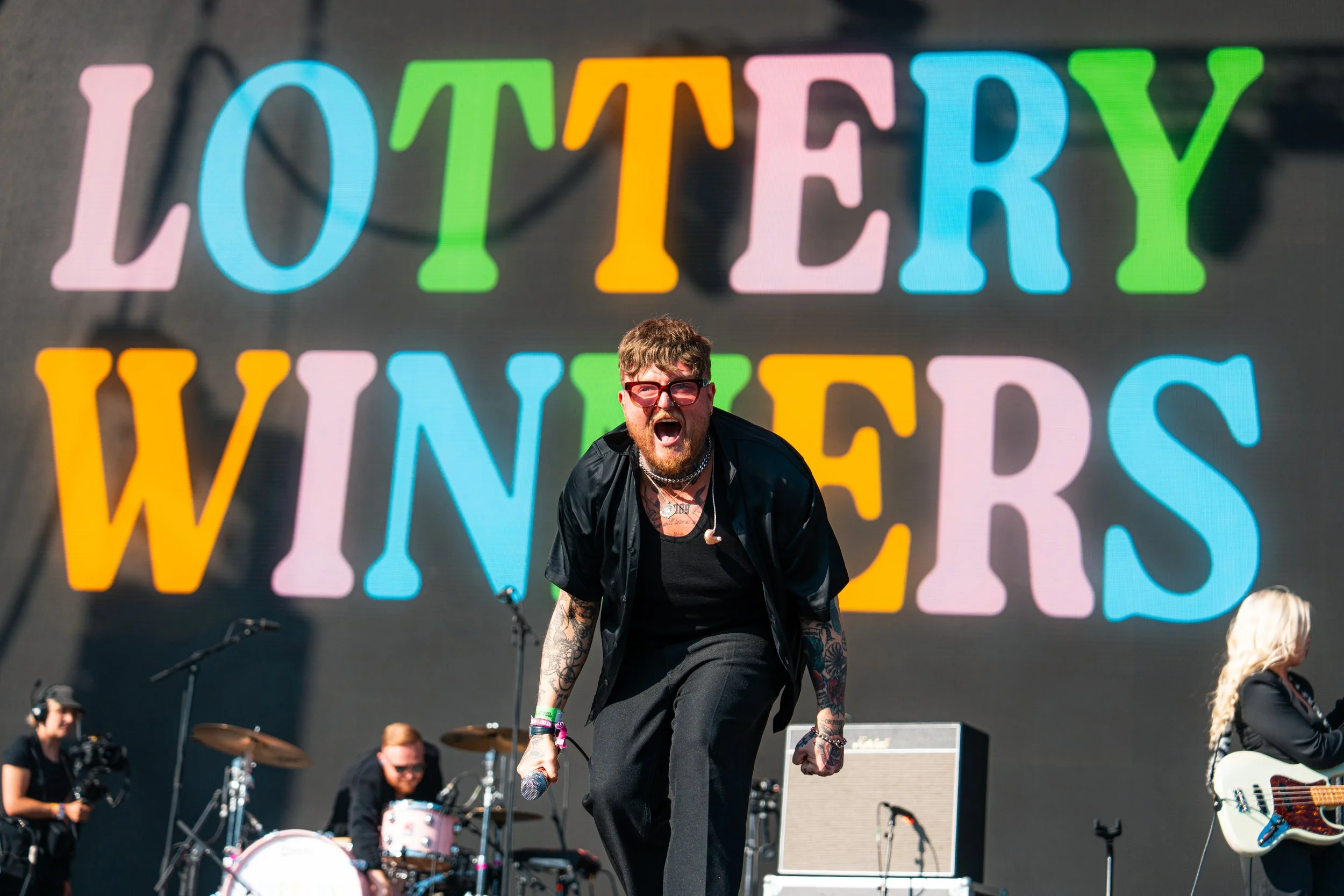 A band performing on stage with a large colorful sign behind them that says 'Lottery Winners'. The lead singer, a man with tattoos, glasses, and a beard, is front and center, passionately singing into a microphone. The Lottery Winners