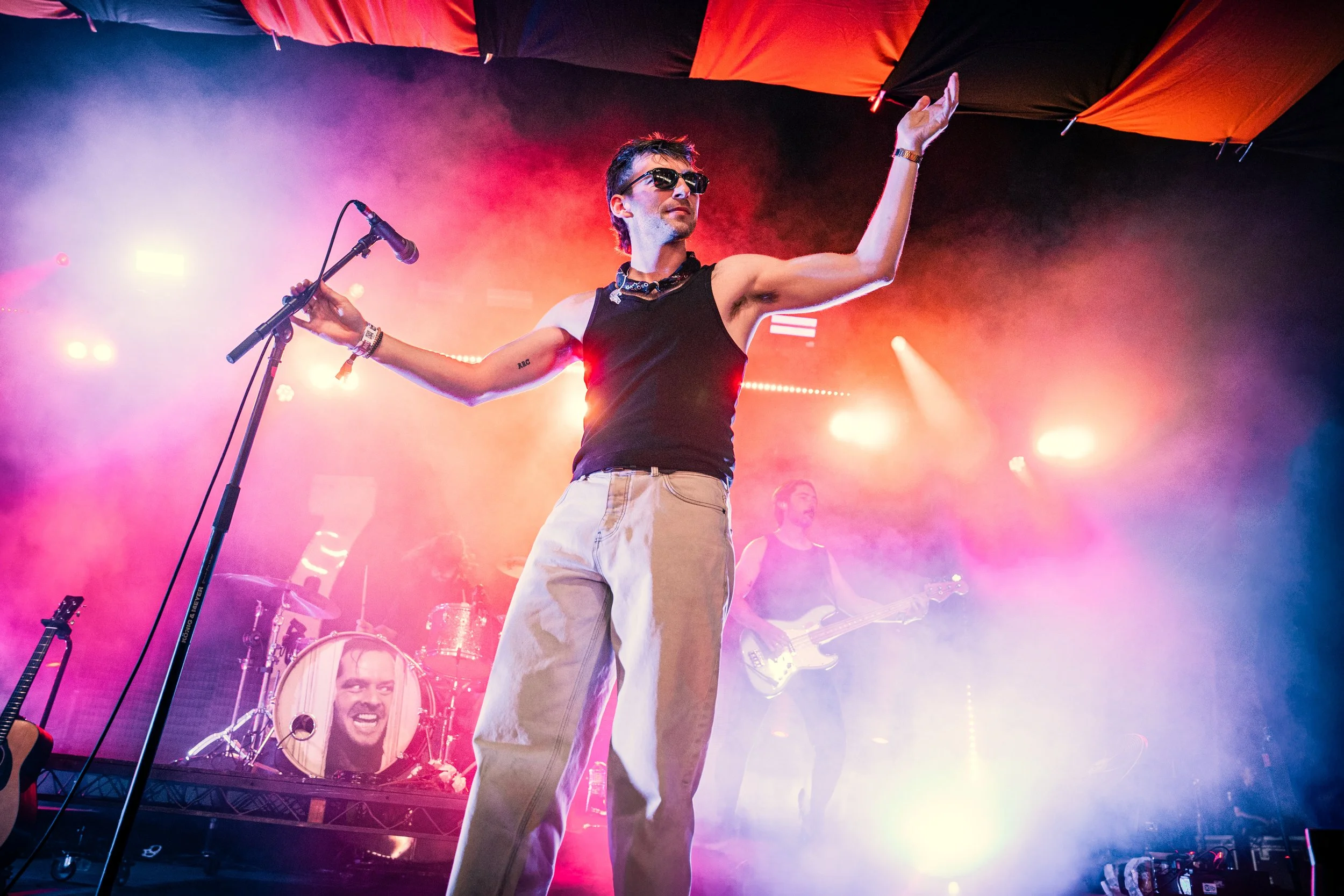 Person performing on stage with microphone, wearing sunglasses, black tank top, and beige pants, with band members playing instruments in the background, amid colorful stage lighting and fog.