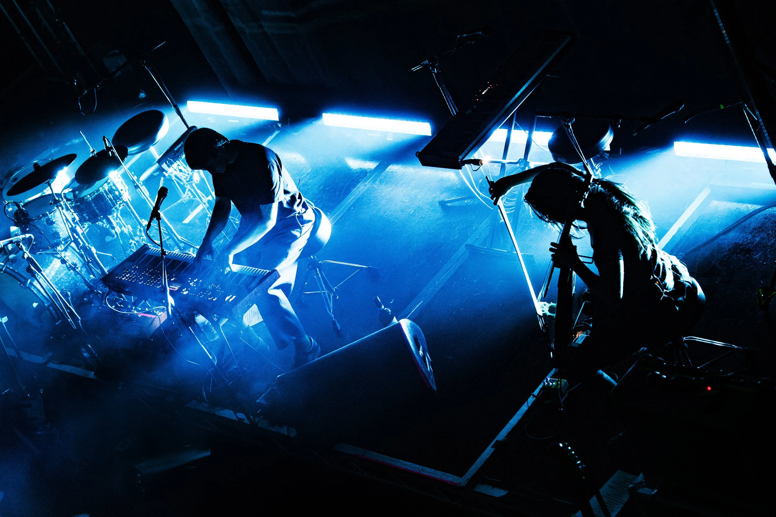 Two musicians performing on stage with electronic equipment, illuminated by blue stage lighting, one adjusting sound gear and the other playing keyboard.