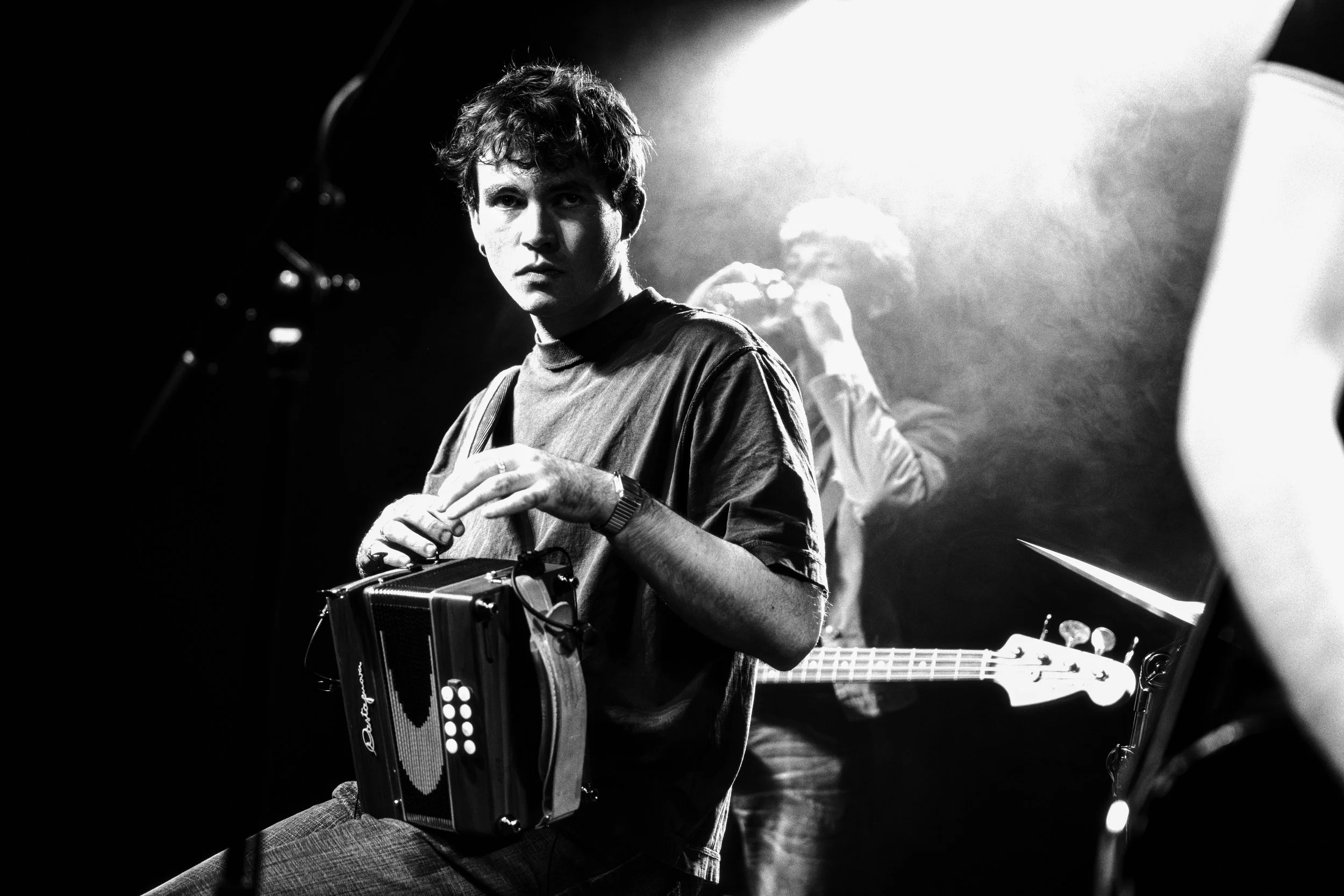 A young man with curly hair sits holding a small keyboard or electronic instrument, with a person in the background playing a bass guitar. The scene is dimly lit with high contrast black and white photography.