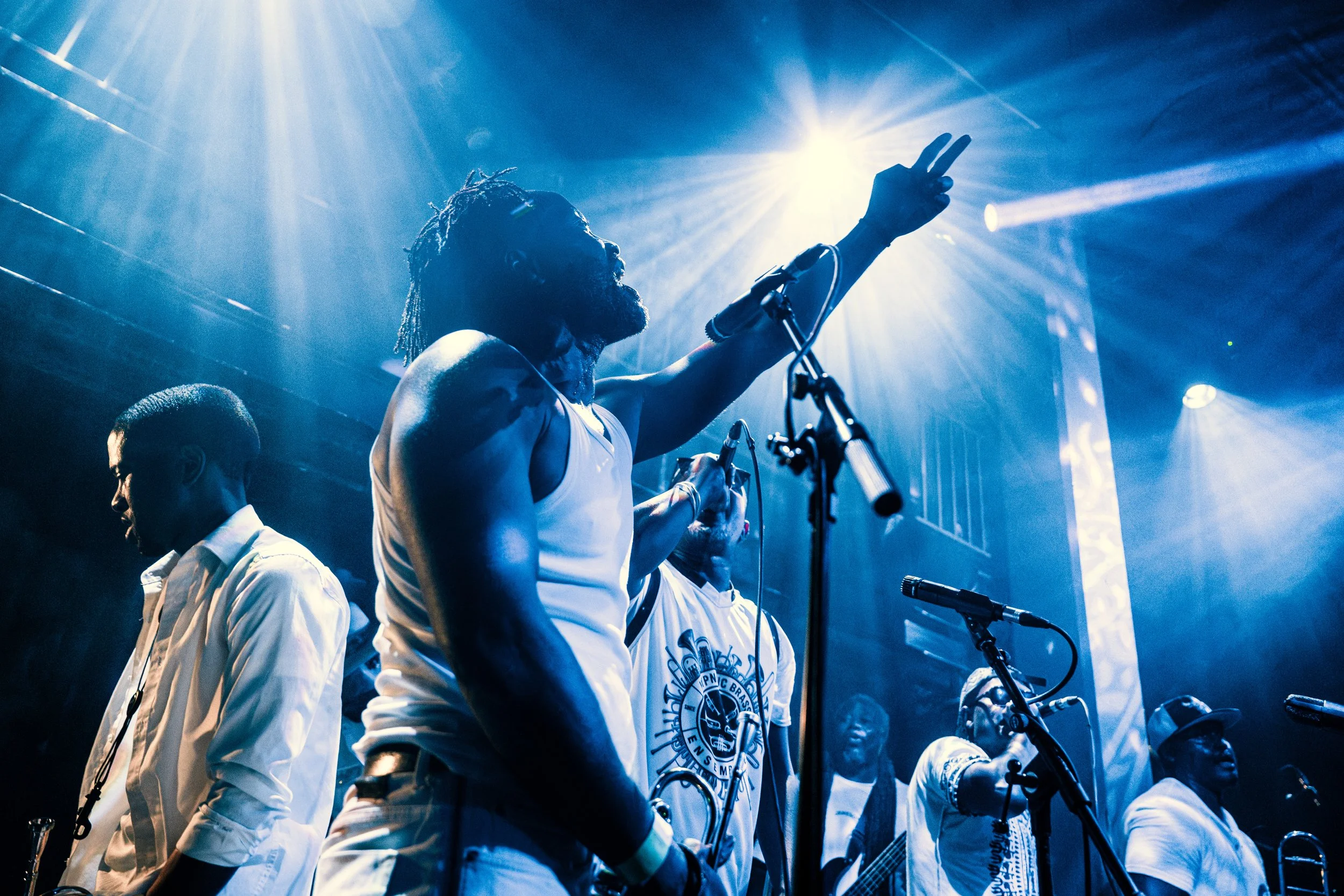 Musicians performing on stage with blue lighting, one man with dreadlocks raising a hand, wearing a white tank top, and others playing instruments and singing.