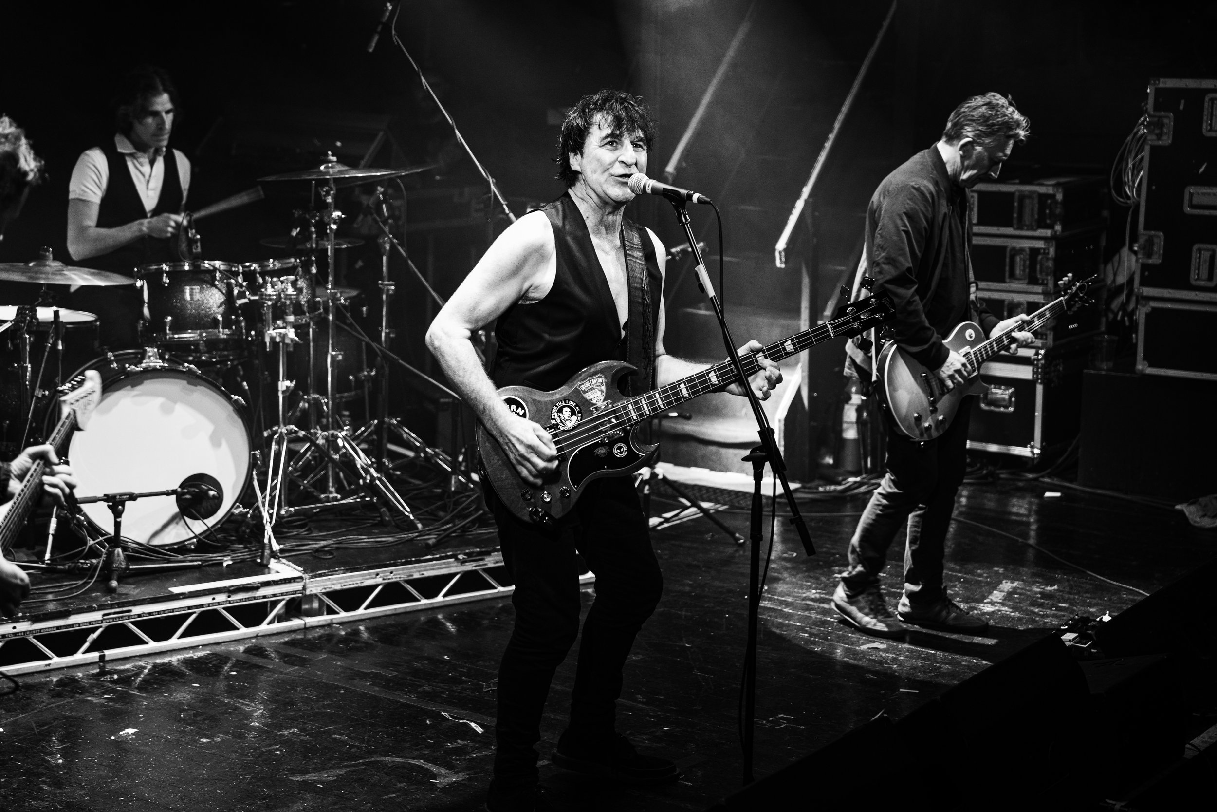 Black and white photo of a band performing on stage with a drummer, guitarist, and bassist.