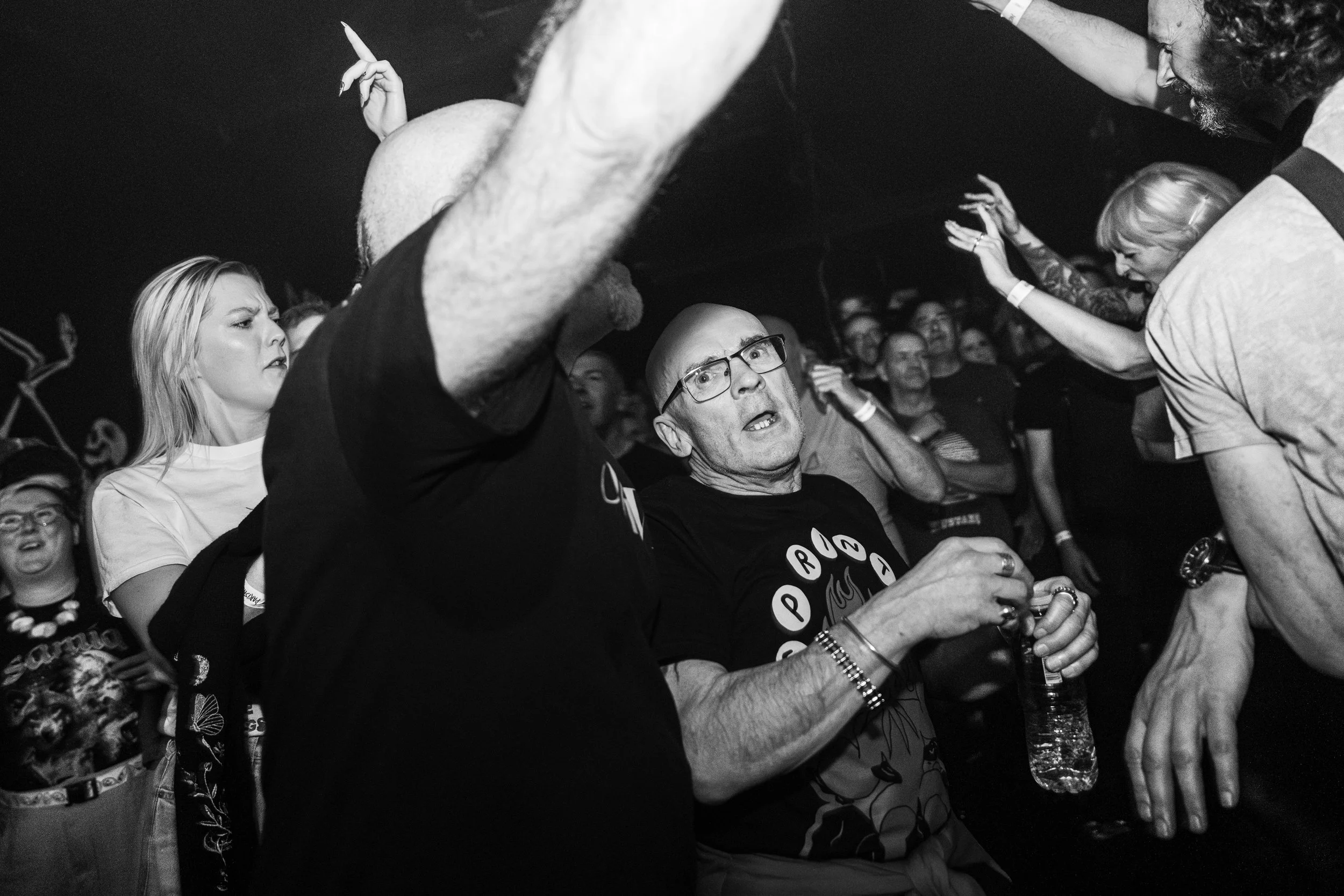 Black and white photo of people at a concert or event. A man with glasses and a tattooed arm appears surprised or upset, holding a water bottle. Surrounding people look engaged with some raising their hands or talking.