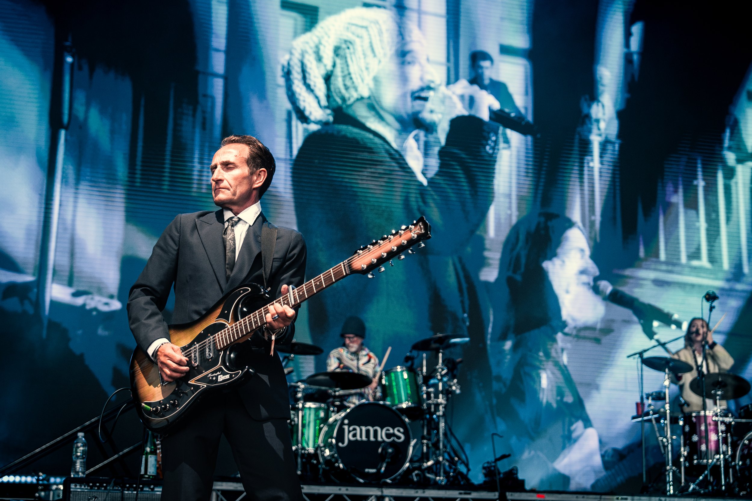 A band performing on stage, featuring a man in a suit playing an electric guitar, a drummer, and a female singer, with a large screen behind them displaying a close-up of a woman with a beanie singing.