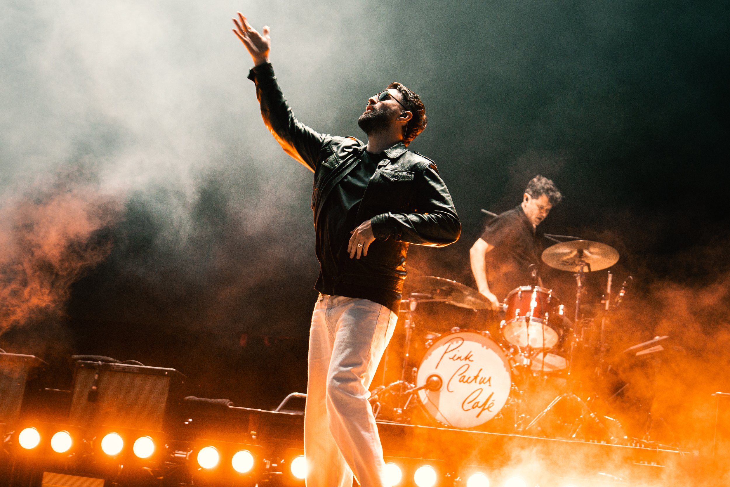 A man wearing sunglasses and a black leather jacket performs on stage with a microphone, with a drummer in the background at Pink Cactus Café. The stage is illuminated with orange lights and smoke.