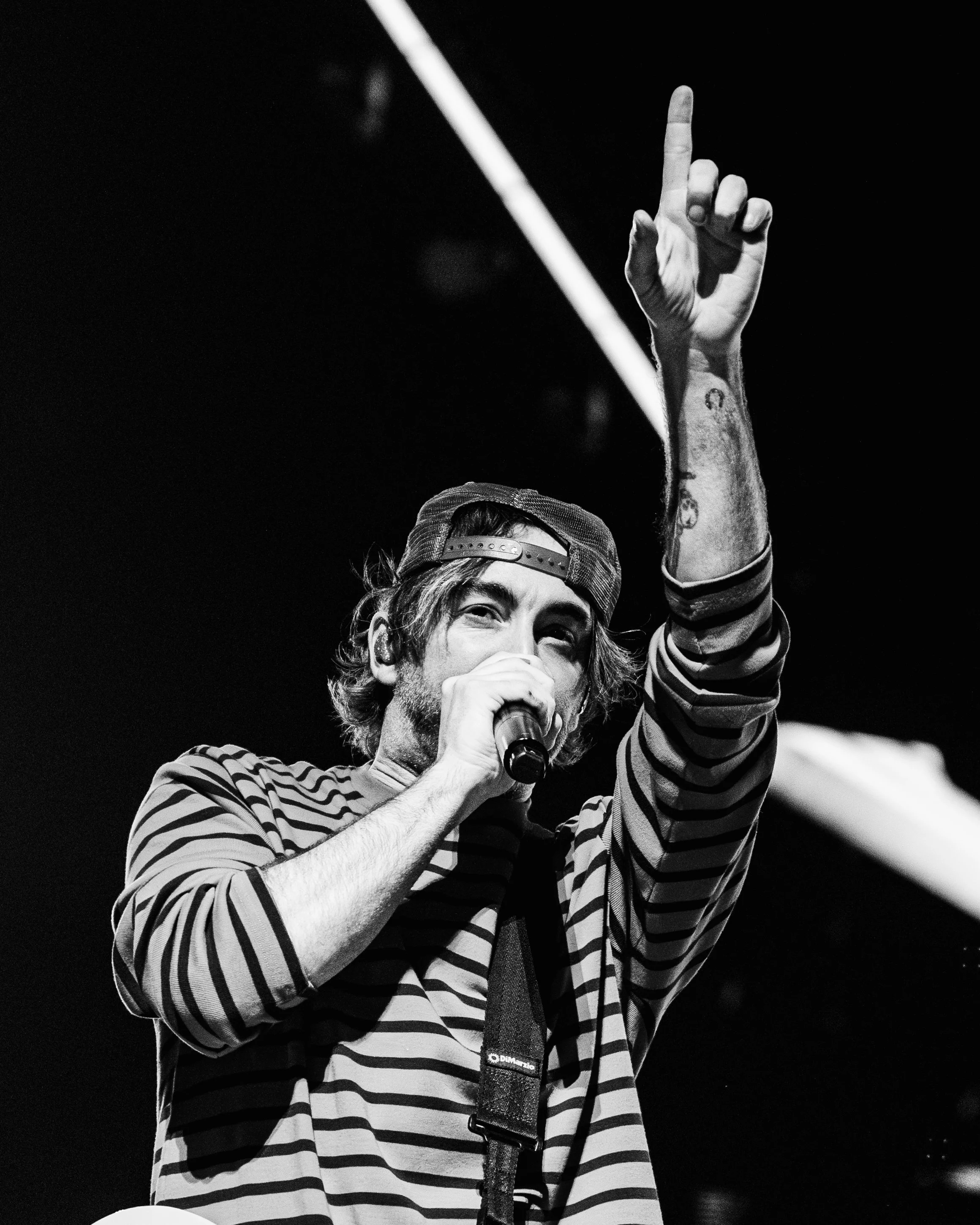 A male performer with long hair, wearing a striped shirt and a backward cap, holding a microphone and raising his index finger during a concert.