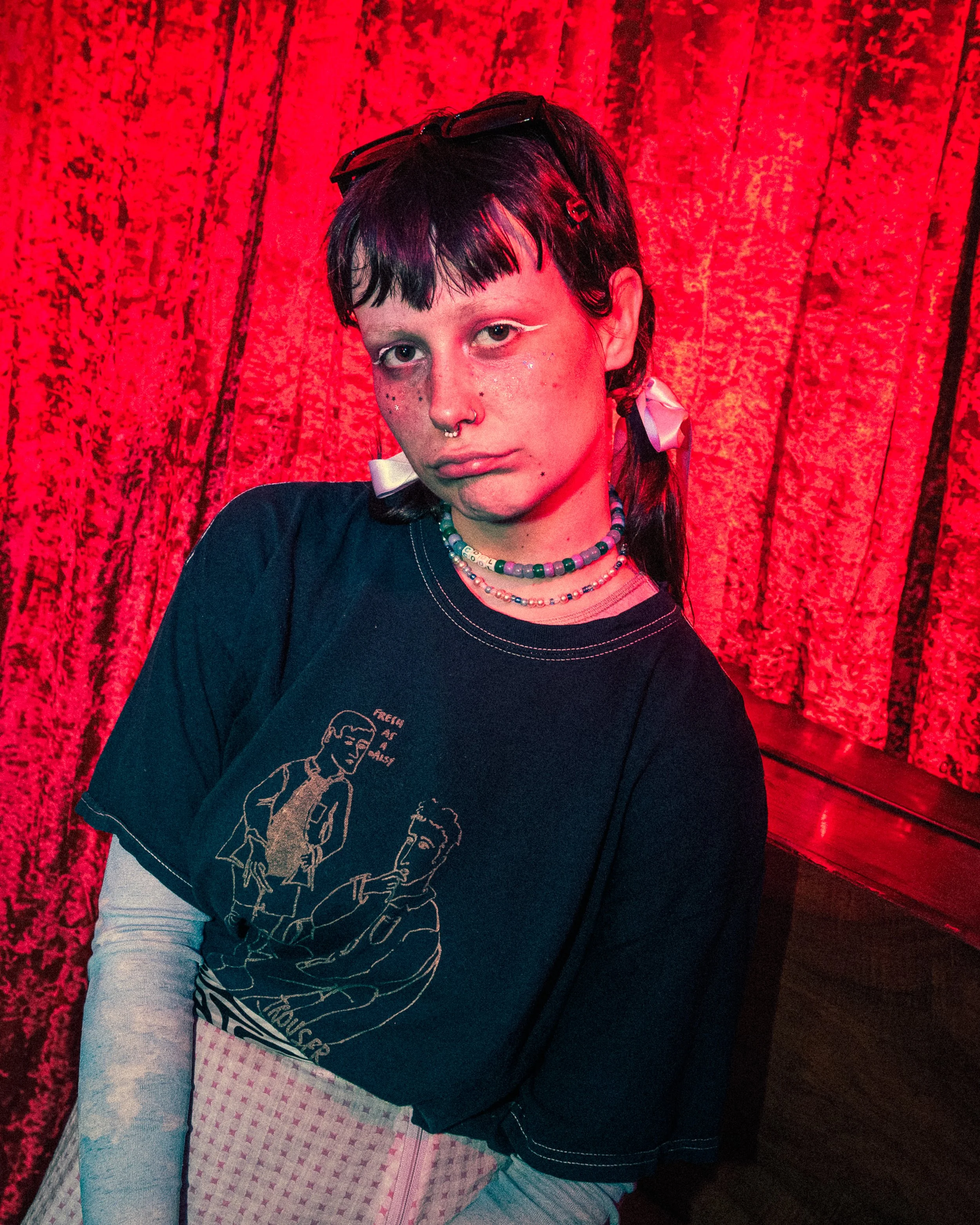 A young woman with short dark hair and tattoos, wearing layered necklaces, a black graphic T-shirt, and a pink checkered skirt, standing in front of a red curtain backdrop.