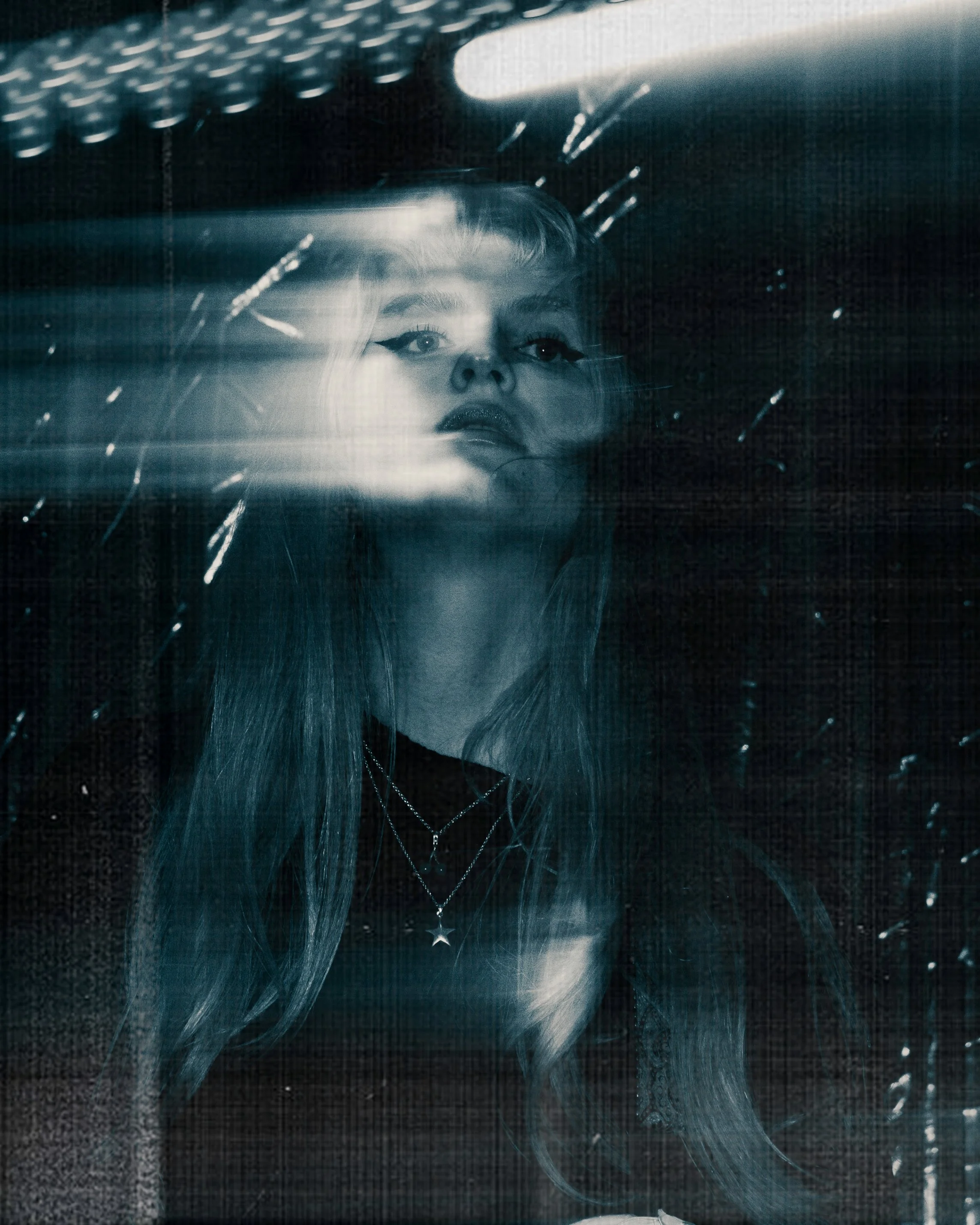 A woman with long hair and makeup looking through a rain-streaked glass window at night.