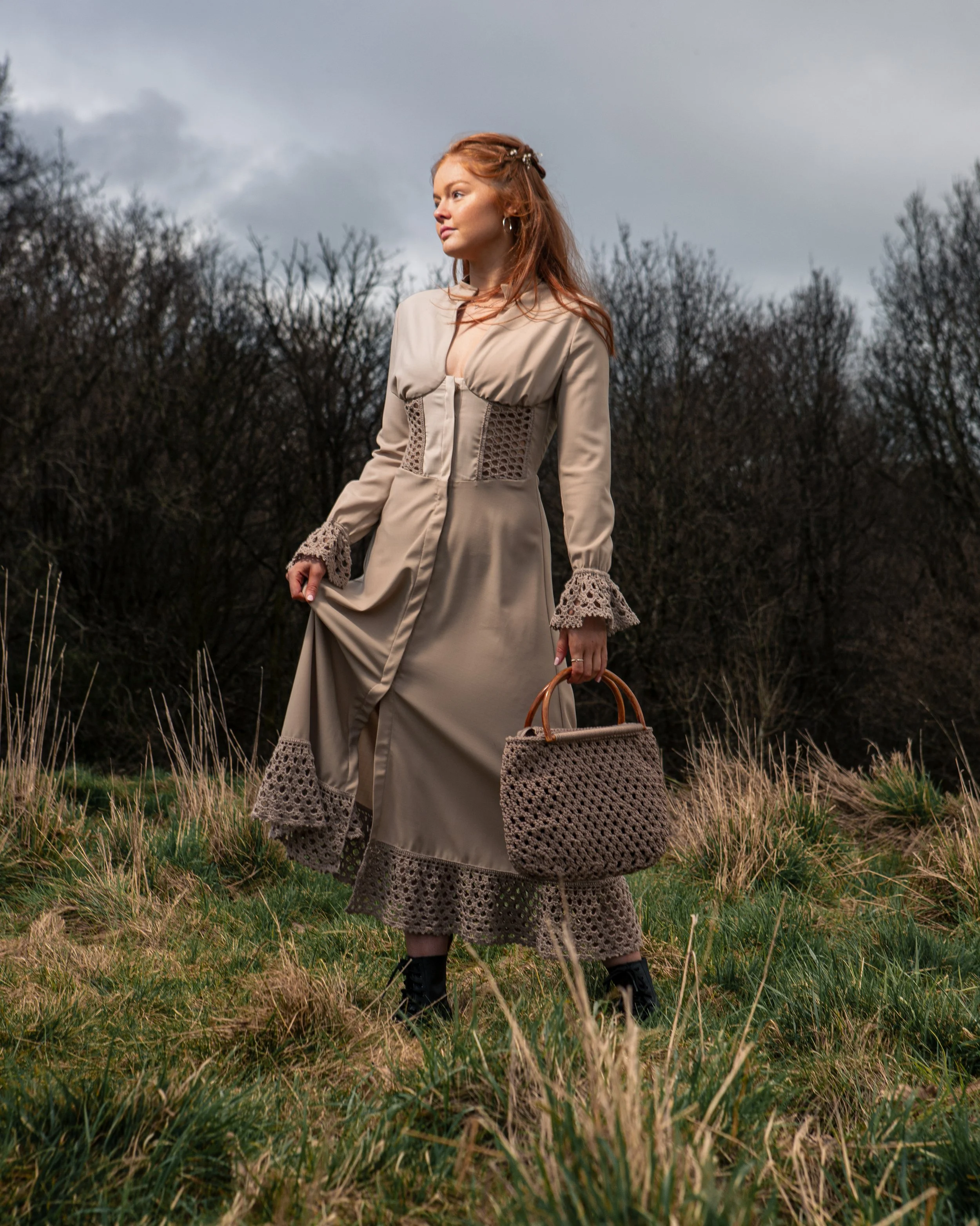 A woman with red hair dressed in a long beige dress with lace accents standing in a grassy field with dark, leafless trees in the background on a cloudy day, holding a woven handbag.