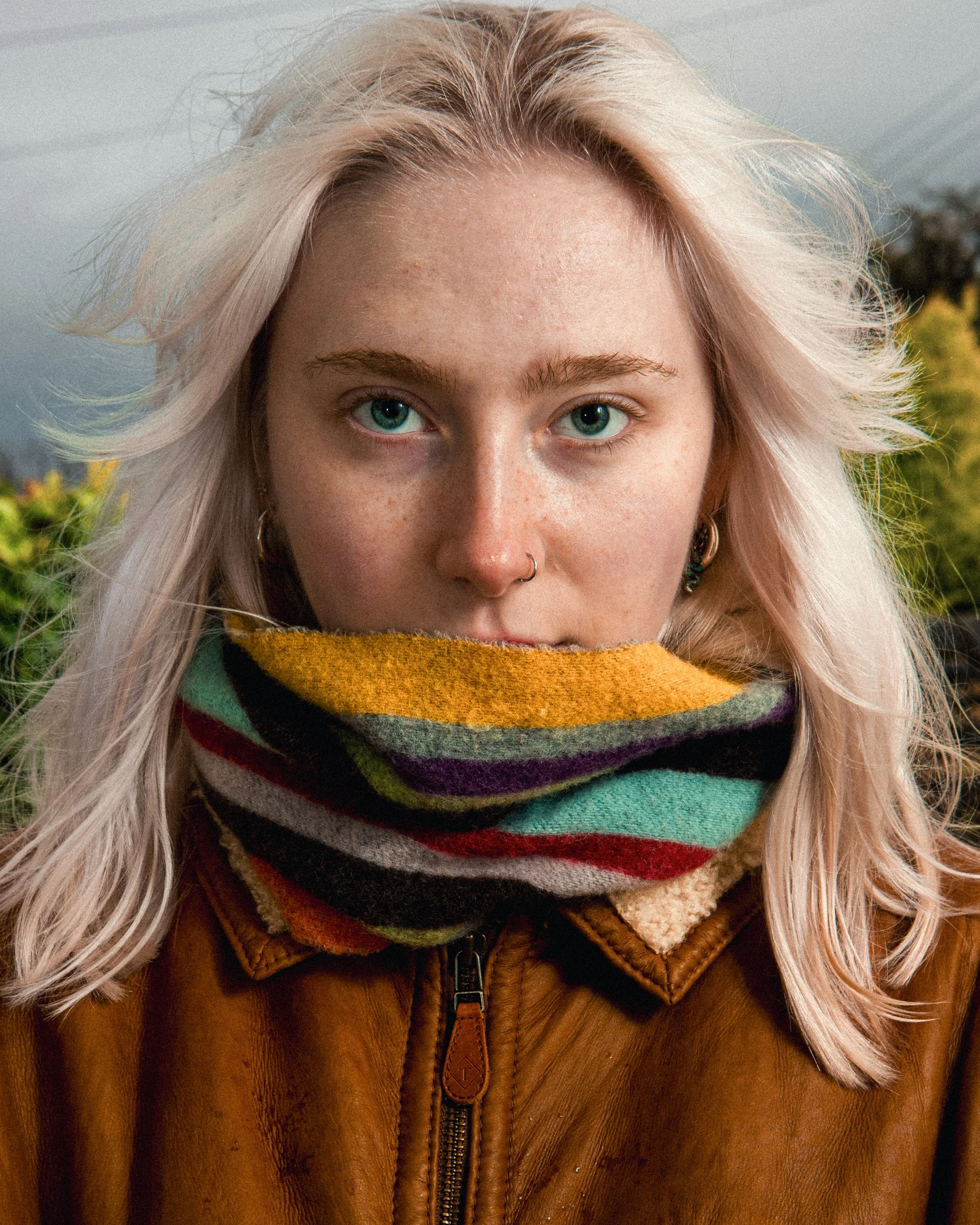 A young woman with blonde hair and blue eyes wearing a colorful striped scarf and a brown leather jacket outdoors.