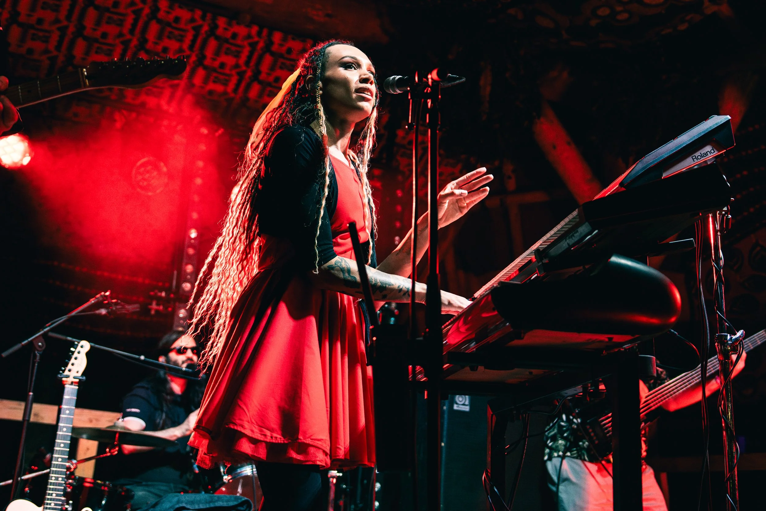 A female musician with long dreadlocks and tattoos, singing into a microphone while playing a keyboard during a live performance, with a guitarist in the background and red stage lighting.
