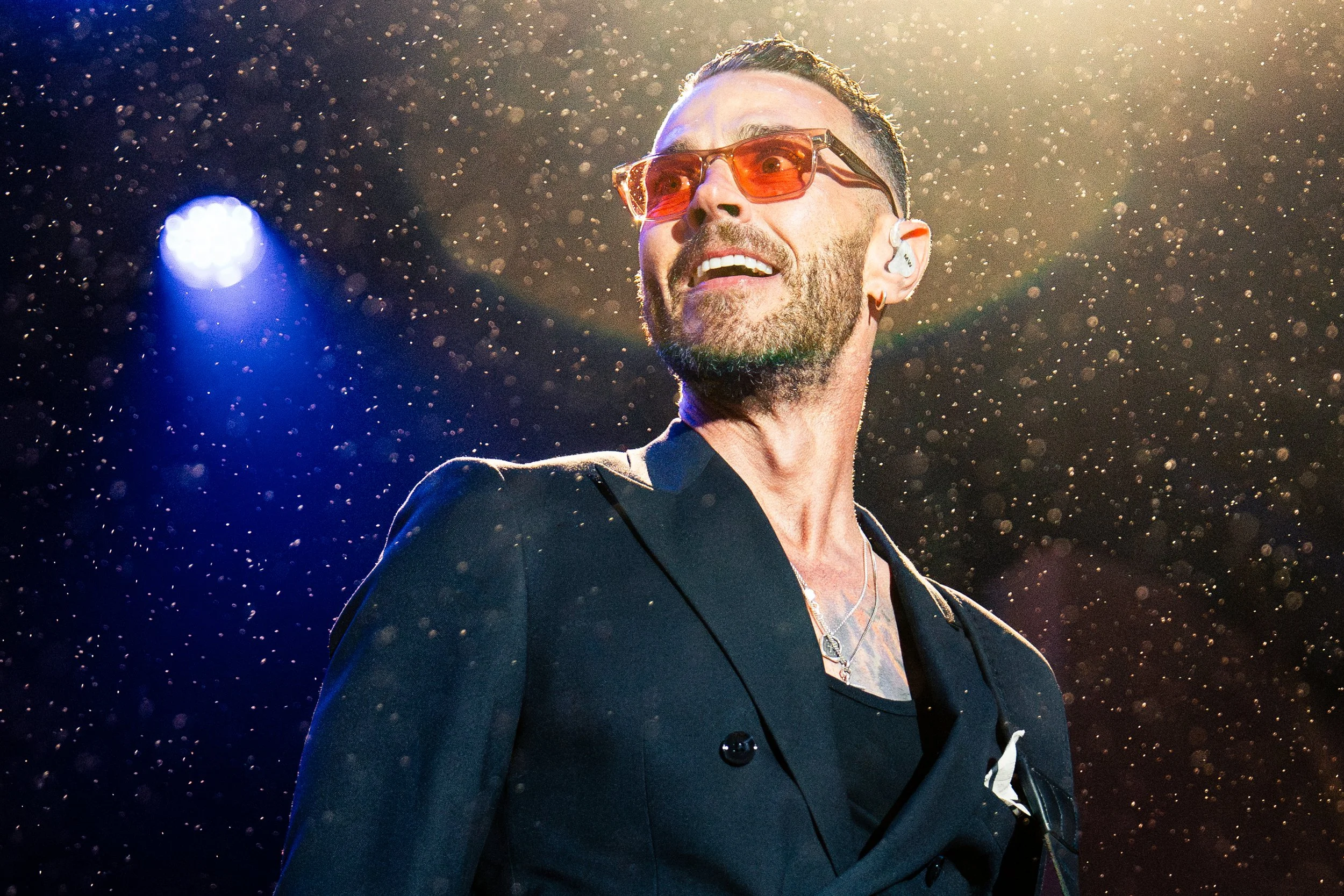 A man on stage during a concert, wearing sunglasses and a black jacket, illuminated by stage lights with water droplets in the air.