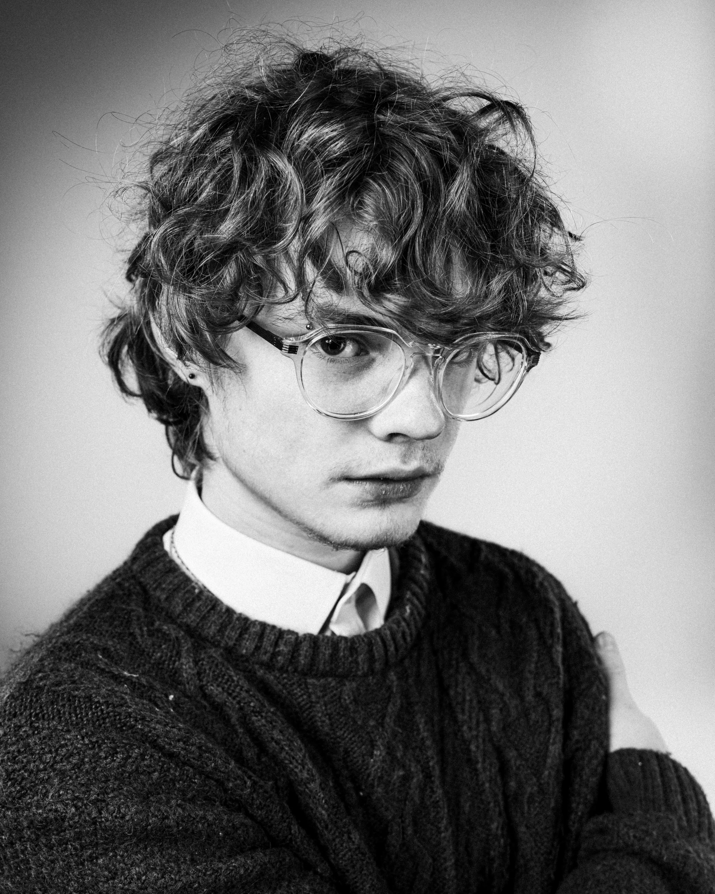 Black and white portrait of a young man with curly hair wearing large glasses, a white shirt, and a dark knitted sweater, looking at the camera with a serious expression.