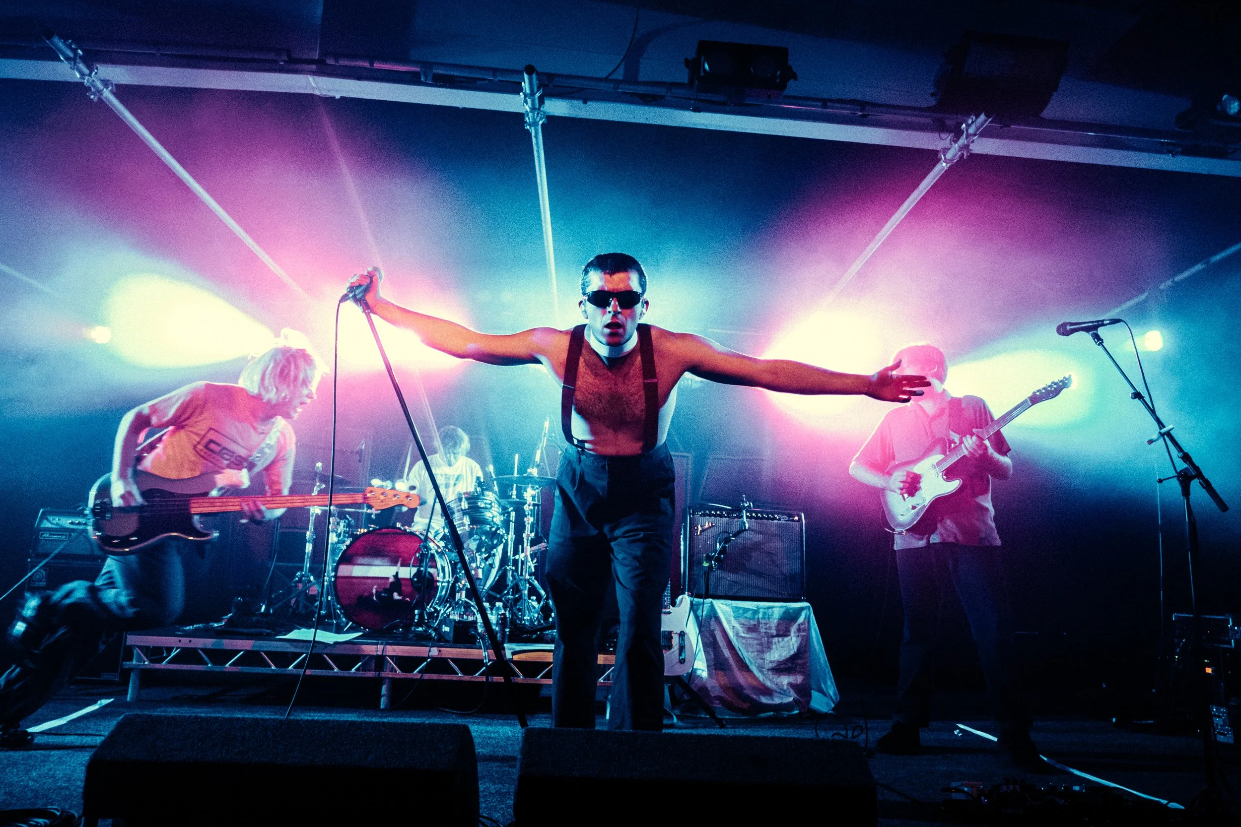 A band performing on stage with colorful lights, featuring a shirtless lead singer wearing sunglasses, holding a microphone, and playing guitar, with other band members playing bass and drums.