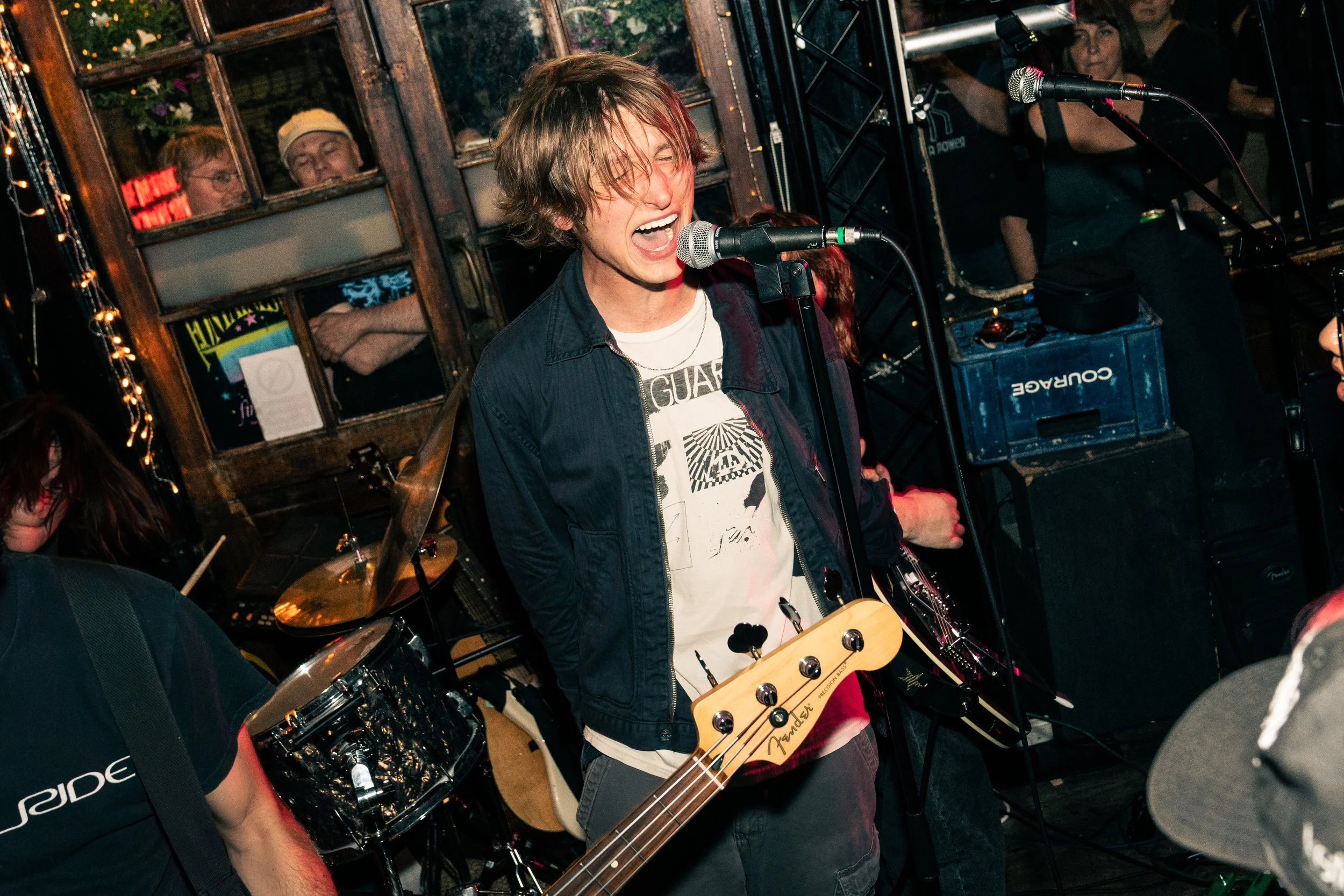 A musician passionately singing into a microphone while playing an electric bass guitar, with an audience in the background at a lively indoor concert.