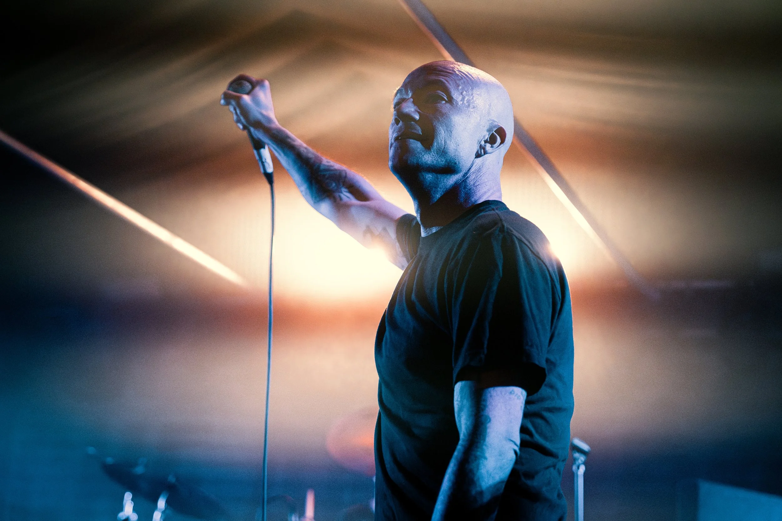 A bald man with tattoos on his arms holding a microphone on stage, illuminated by colorful stage lights.