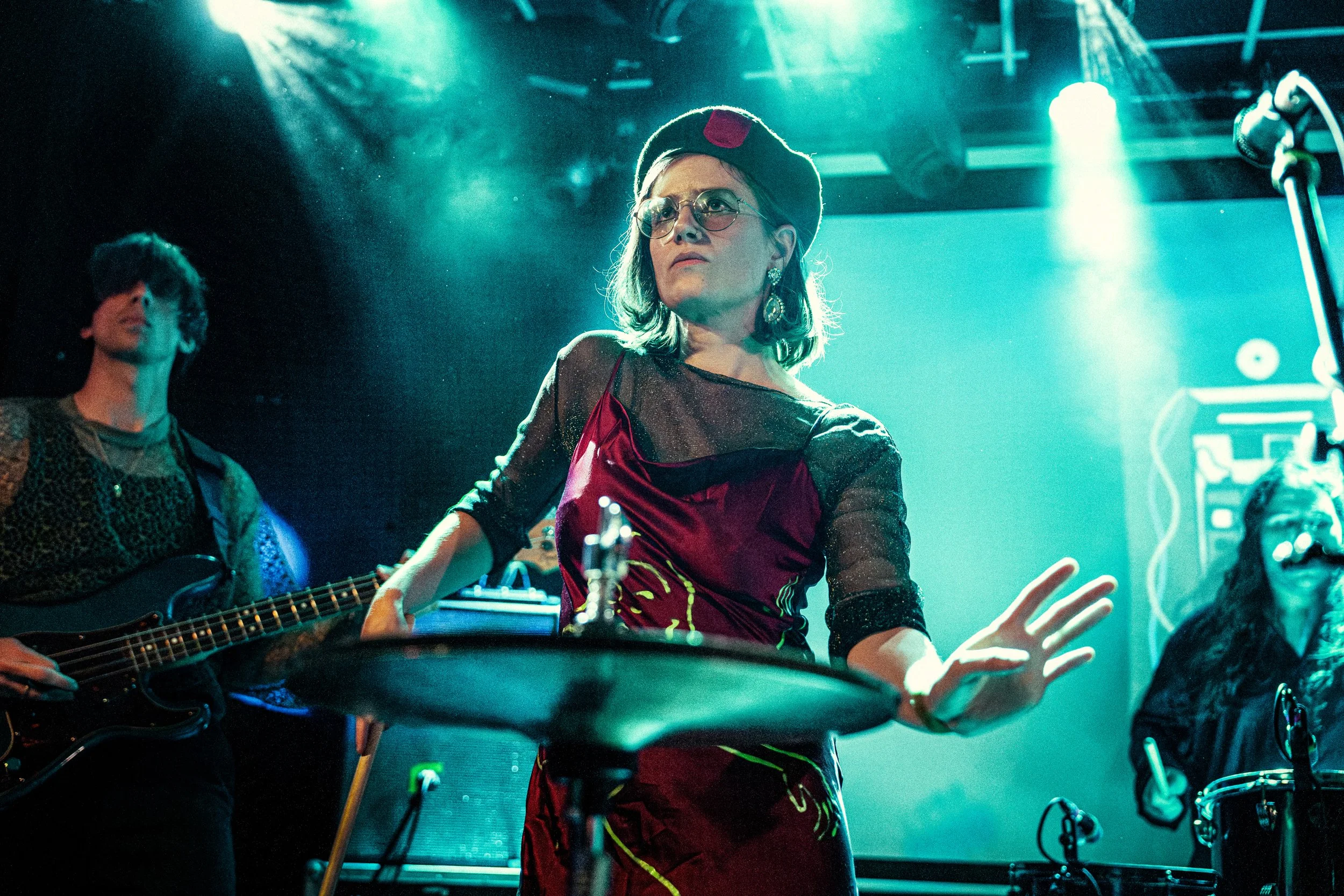 A woman wearing glasses, a black beret, and a red velvet dress performs on stage with a drum kit. She is focused and gesturing with her right hand while a guitarist stands behind her. The stage is illuminated with blue and green lighting.