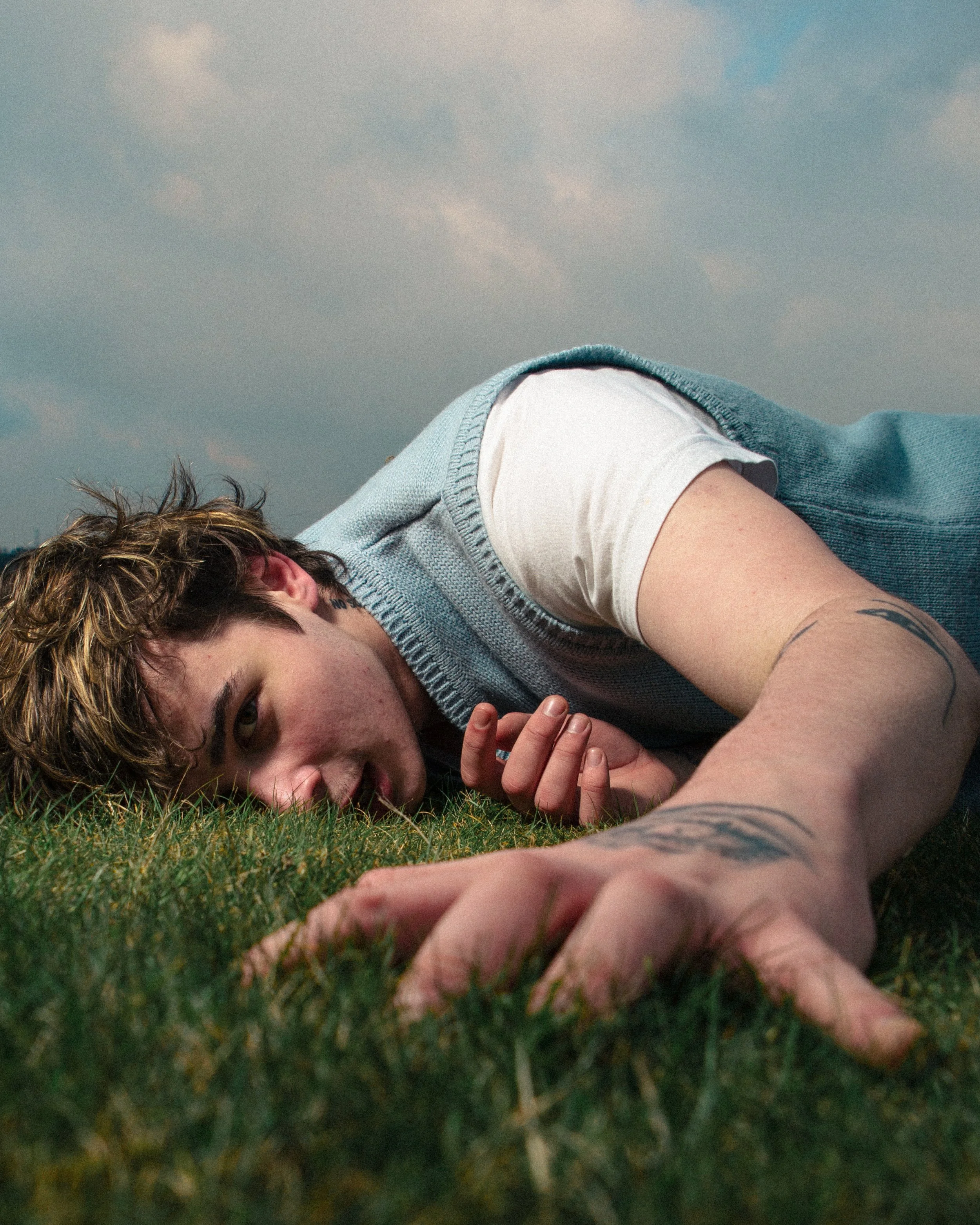 A young man with tattoos in a blue vest and white t-shirt laying on the grass, looking towards the camera, with a partly cloudy sky in the background.