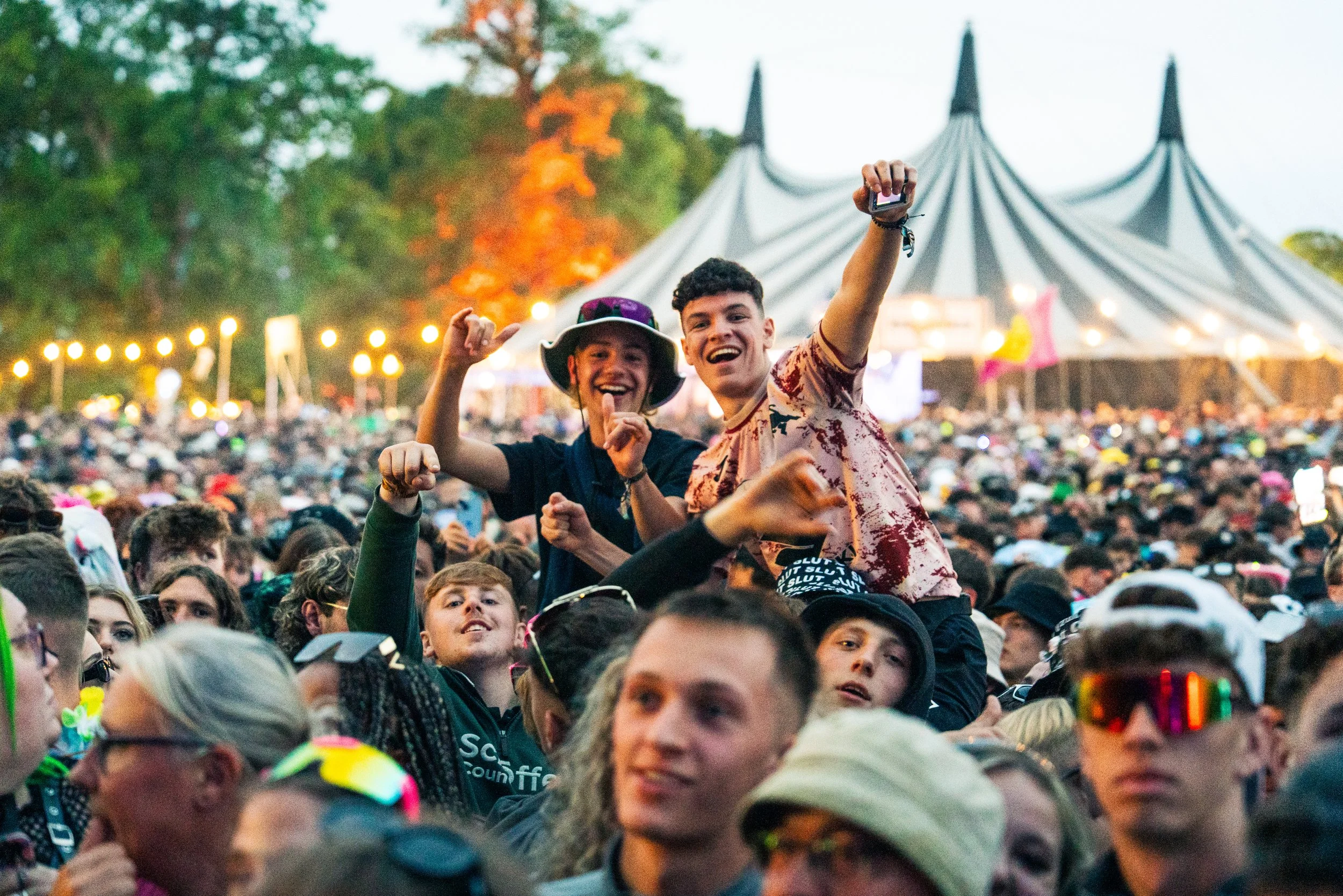 Two young men are smiling and celebrating on the shoulders of the crowd at a large outdoor music festival, with a big tent and trees in the background.