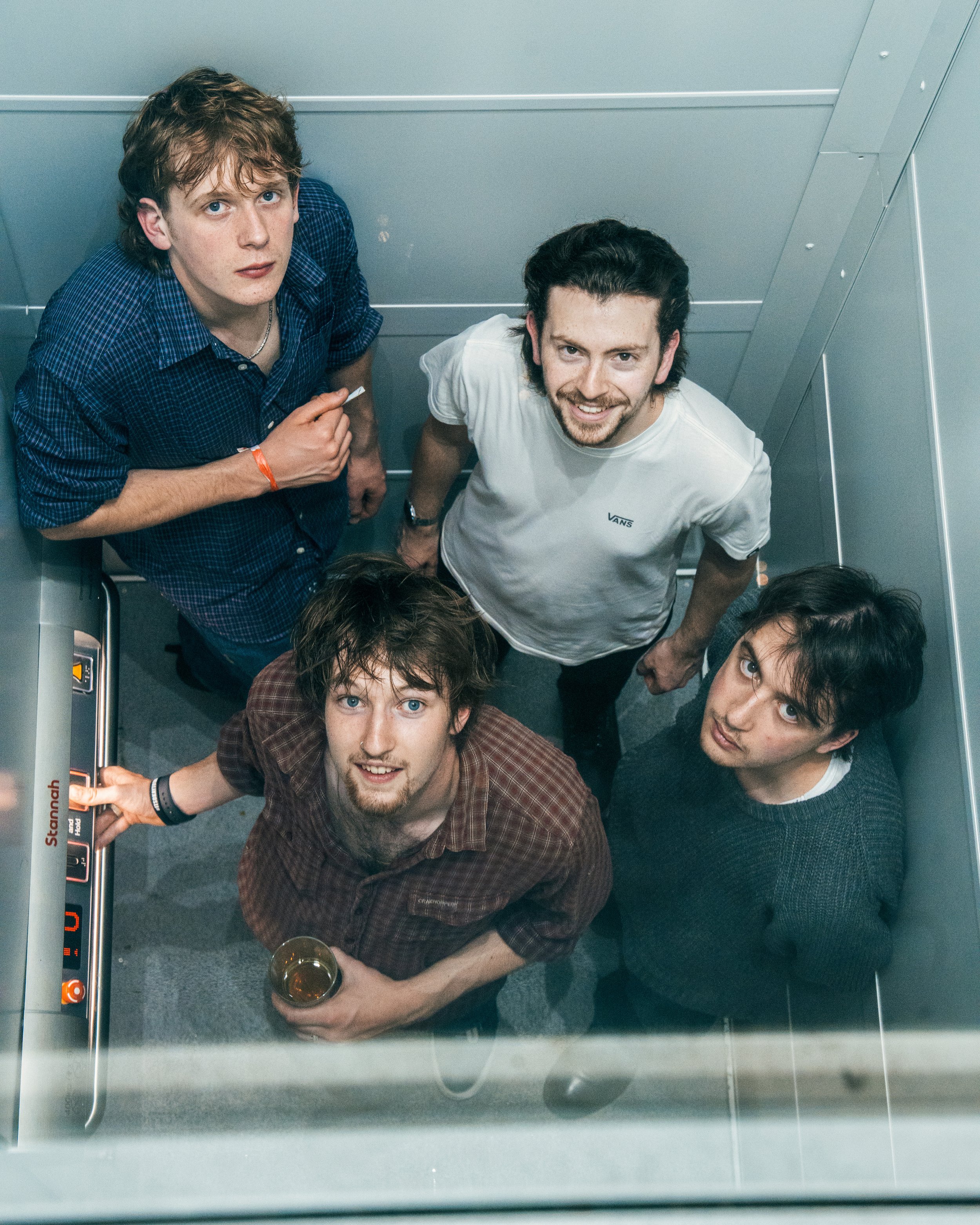 Five young men looking up and smiling inside an elevator, with one holding a drink, and one pressing a button.