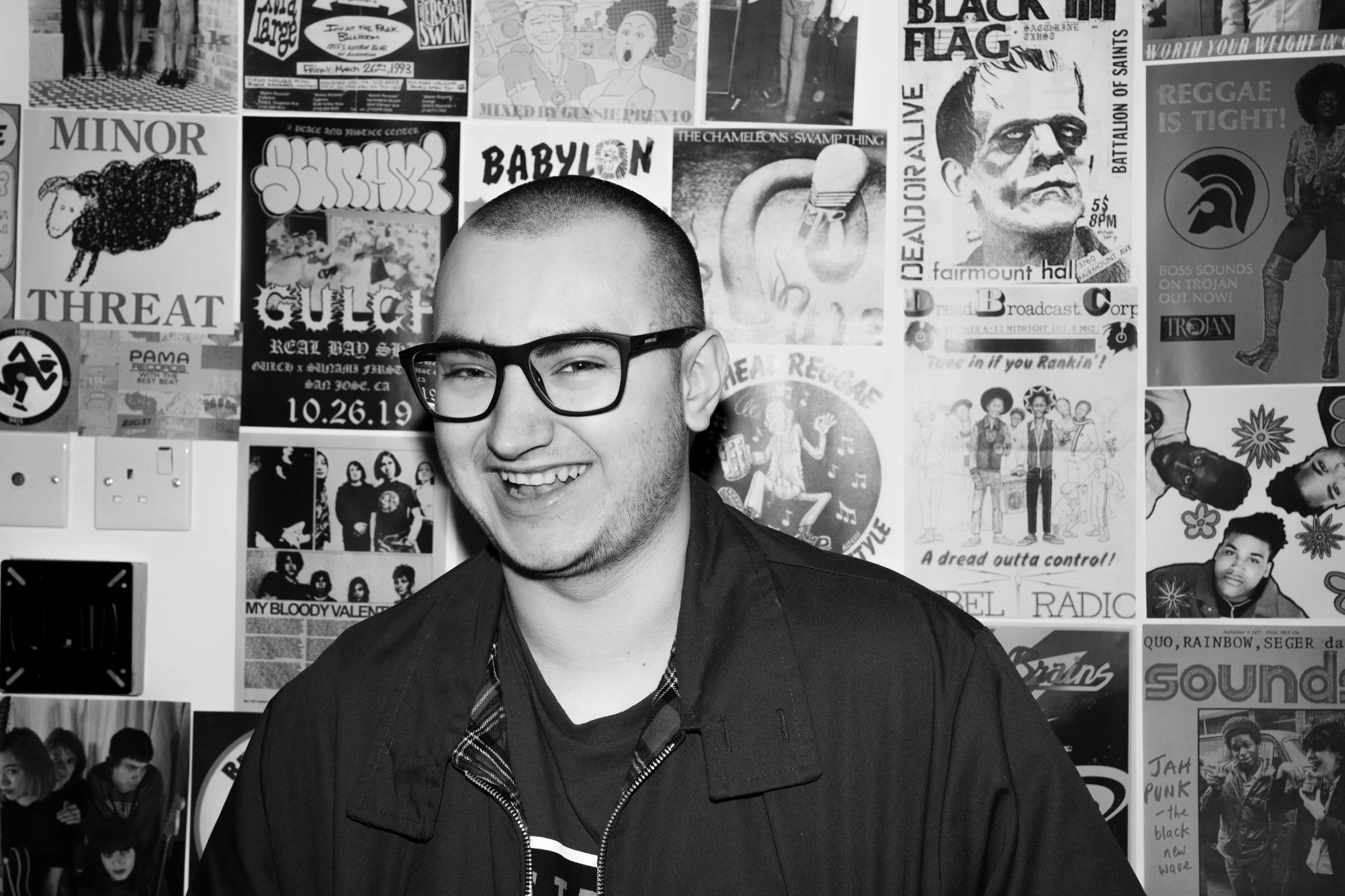 A young man with glasses smiling in front of a wall covered with various posters and album covers.