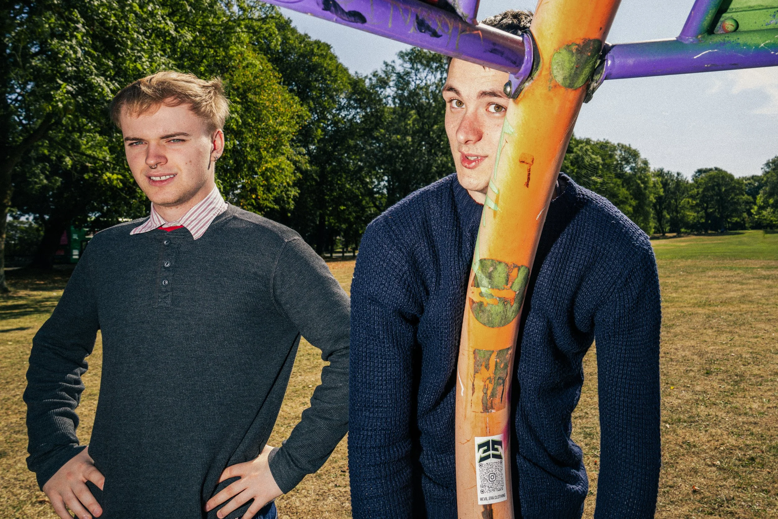 Two young men standing outdoors in a park, one smiling with one hand on his hip, the other holding a skateboard with a colorful deck, both wearing sweaters, trees in the background.