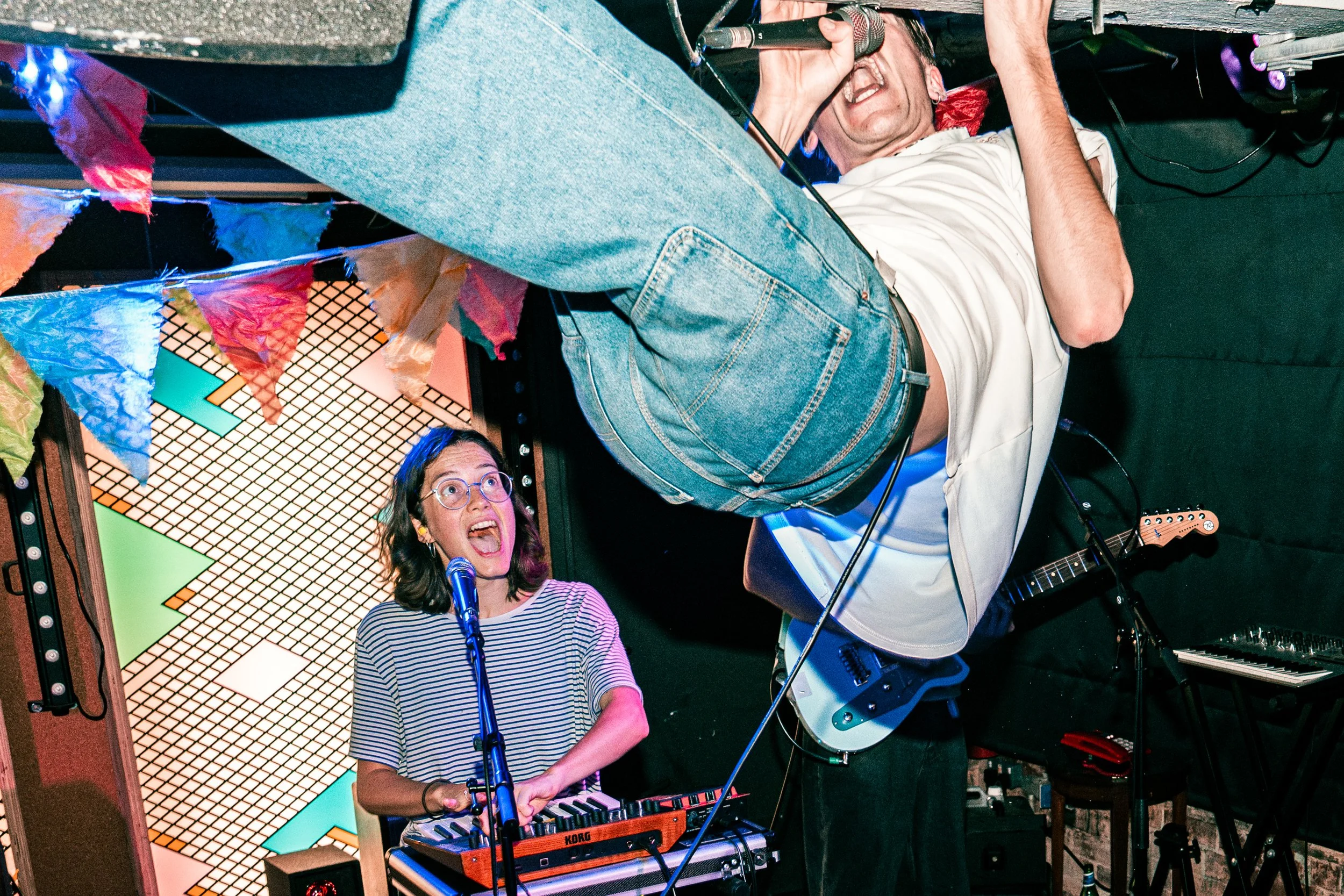 Two musicians performing on stage, one playing a keyboard and singing passionately with glasses, and the other gripping a microphone while standing upside down, wearing jeans and a white t-shirt, under colorful decorations and stage lighting.