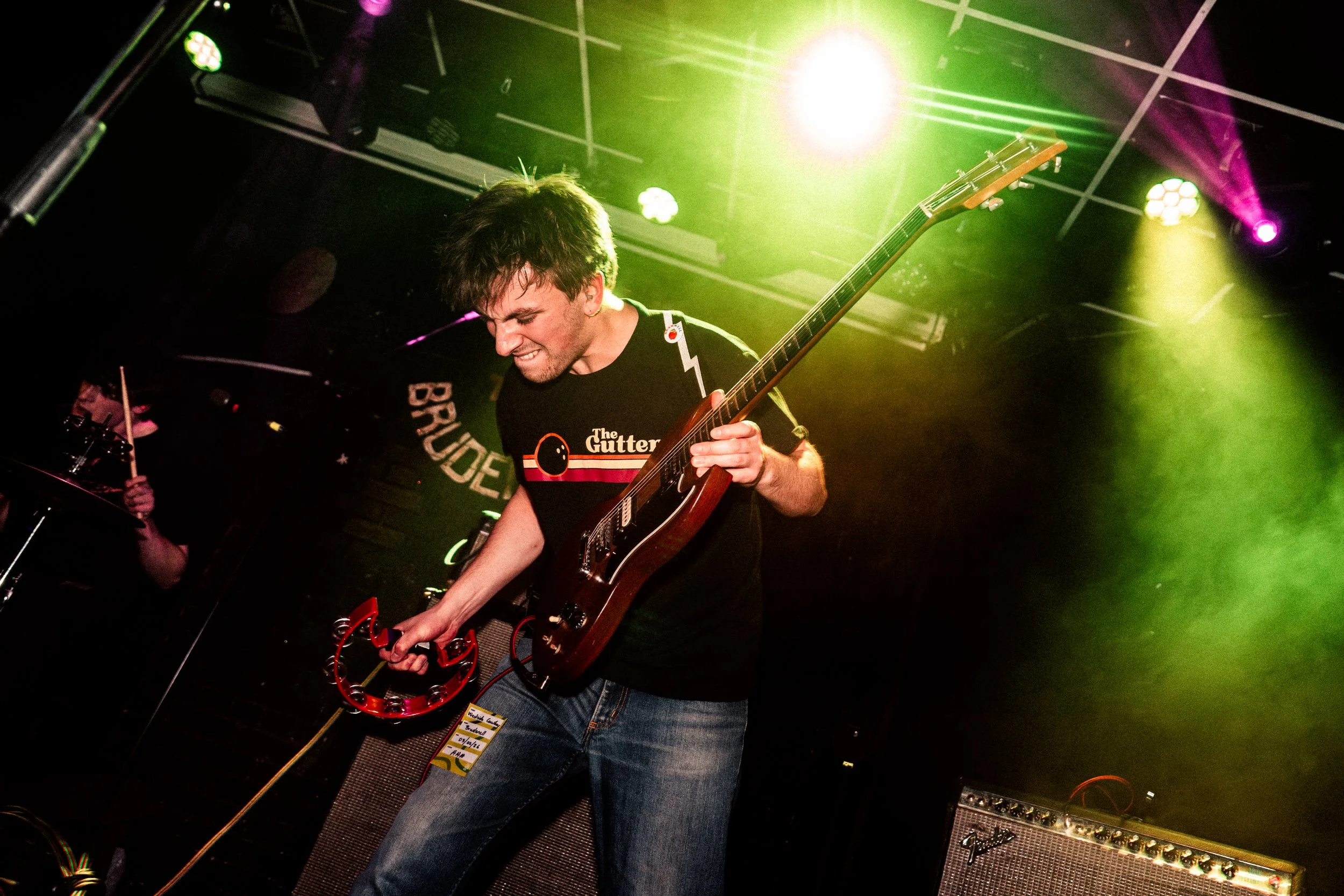 A young man playing an electric guitar on stage under colorful lights, smiling with eyes closed, wearing a black T-shirt and jeans, with a third musician partially visible in the background.