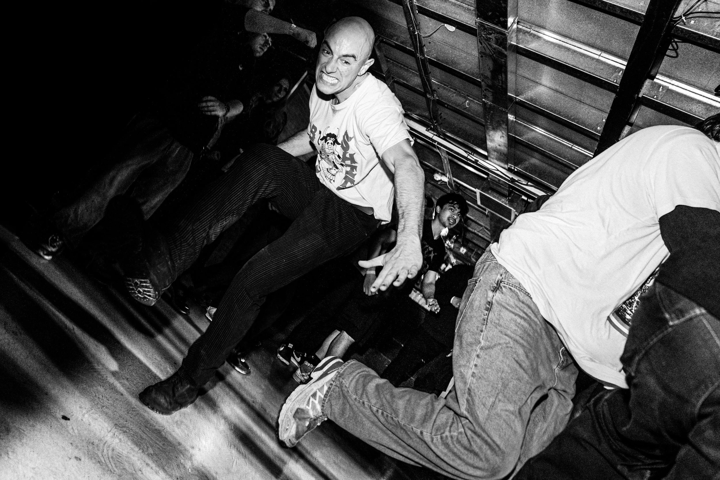 Crowd dancing energetically at a party or concert, with a bald man in a white t-shirt and striped pants in the foreground, making a fierce facial expression.