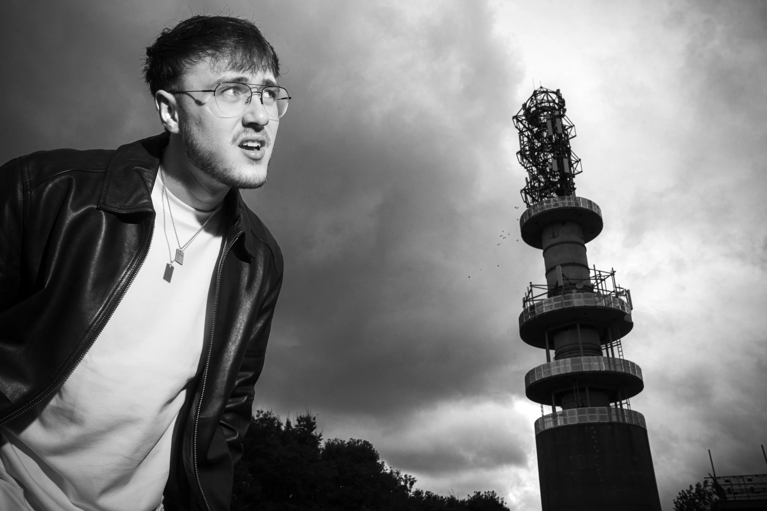 A young man with glasses, a leather jacket, and necklaces poses outdoors against a cloudy sky, with a tall communication tower in the background.