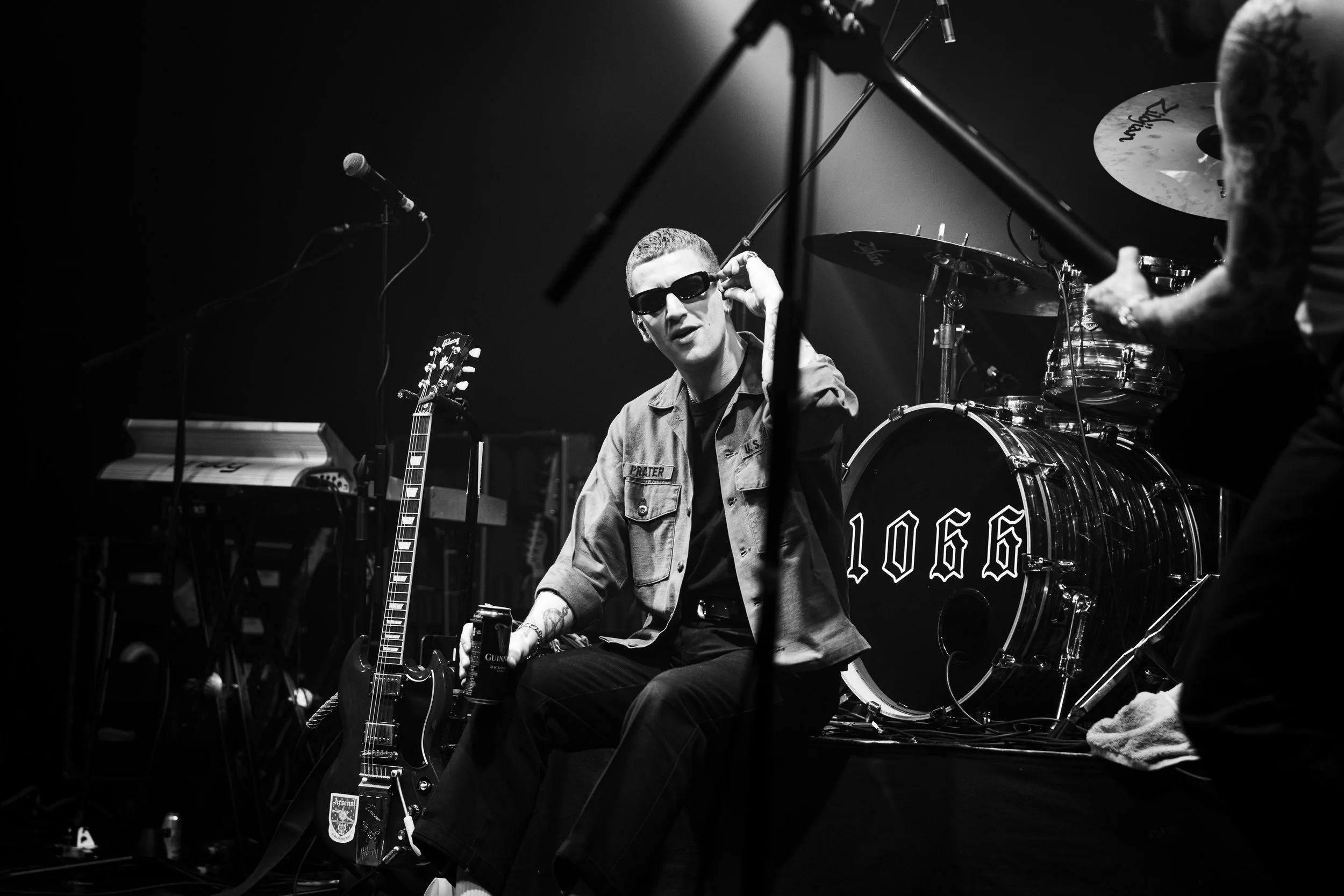 A black and white photo of a musician on stage with sunglasses, holding a soda can, surrounded by musical instruments including a guitar, keyboard, and drums, during a live performance.