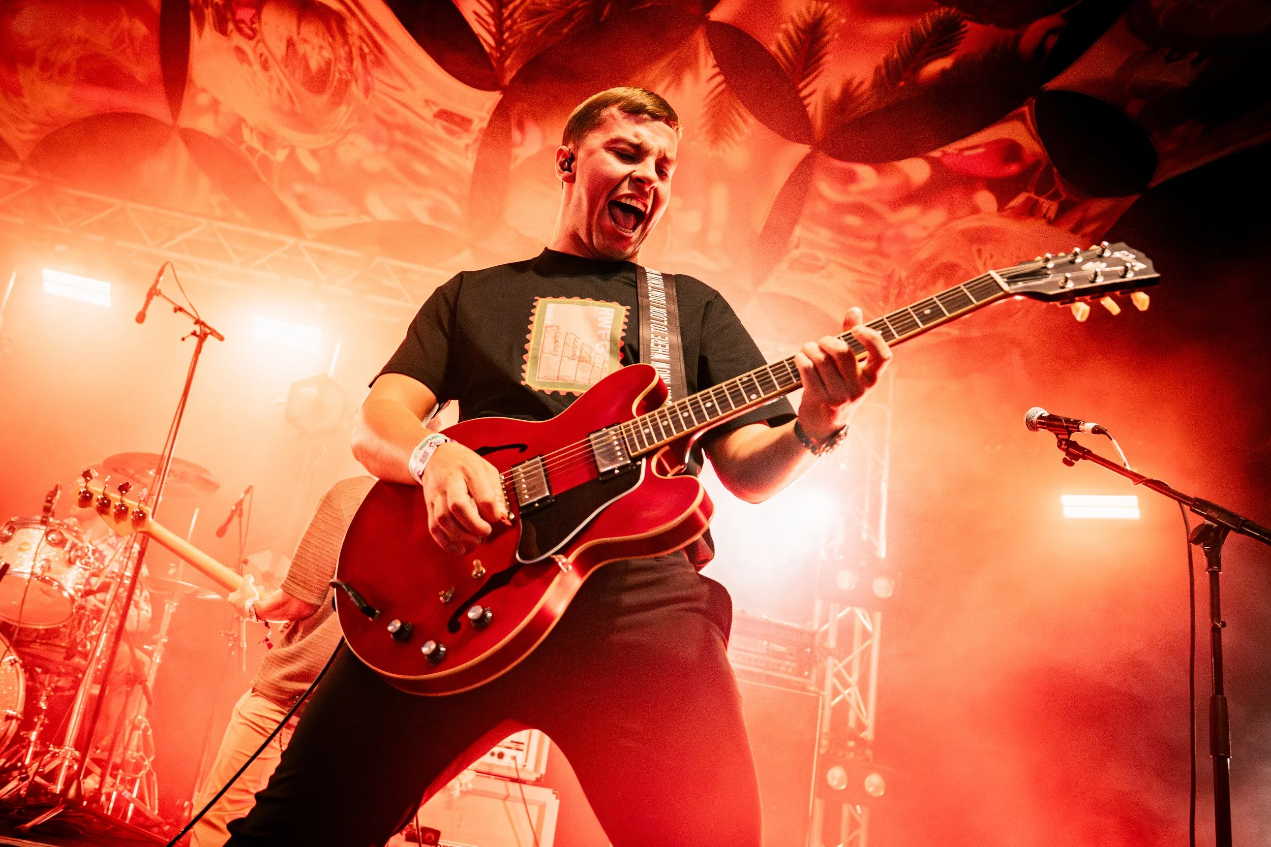 A guitarist passionately playing an electric guitar on stage during a concert, with stage lights and a drum set in the background.