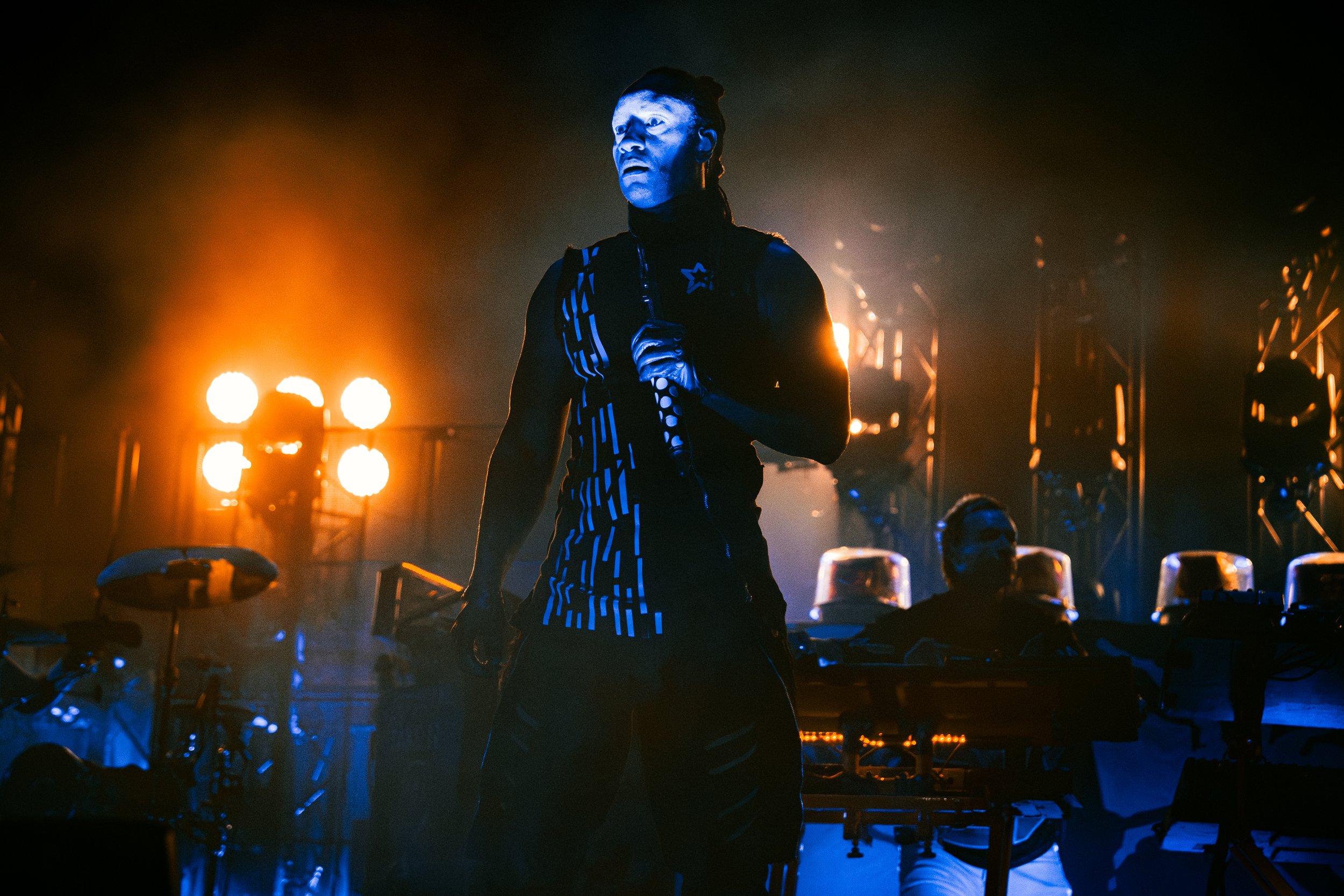 A performer with face paint, wearing a black outfit with blue and white striped accents, holding a microphone stand on a stage illuminated by orange and blue lights, with DJ or music equipment in the background.
