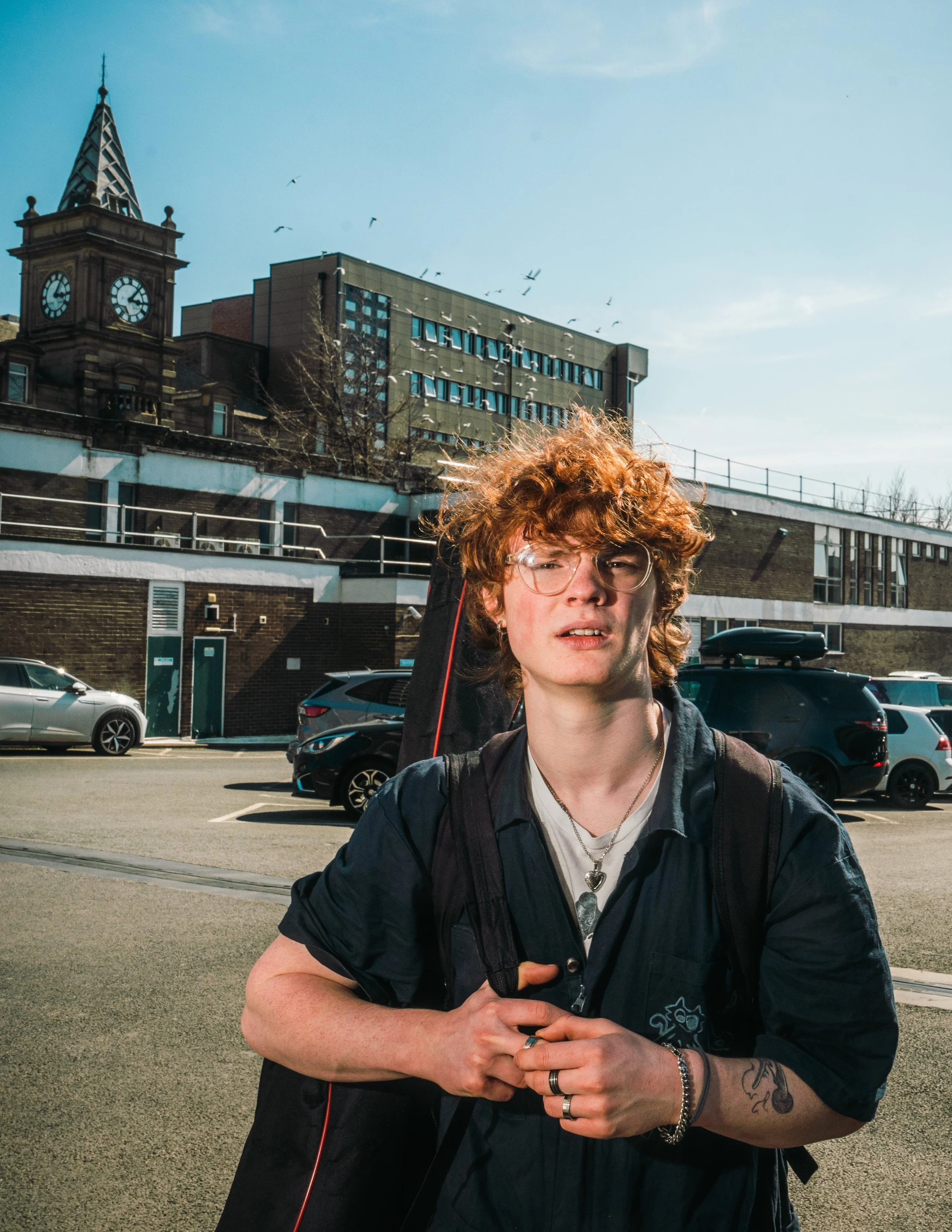 Young man with curly red hair and glasses standing outdoors in an urban area with parked cars, a building with multiple windows, a clock tower, and a clear blue sky in the background.