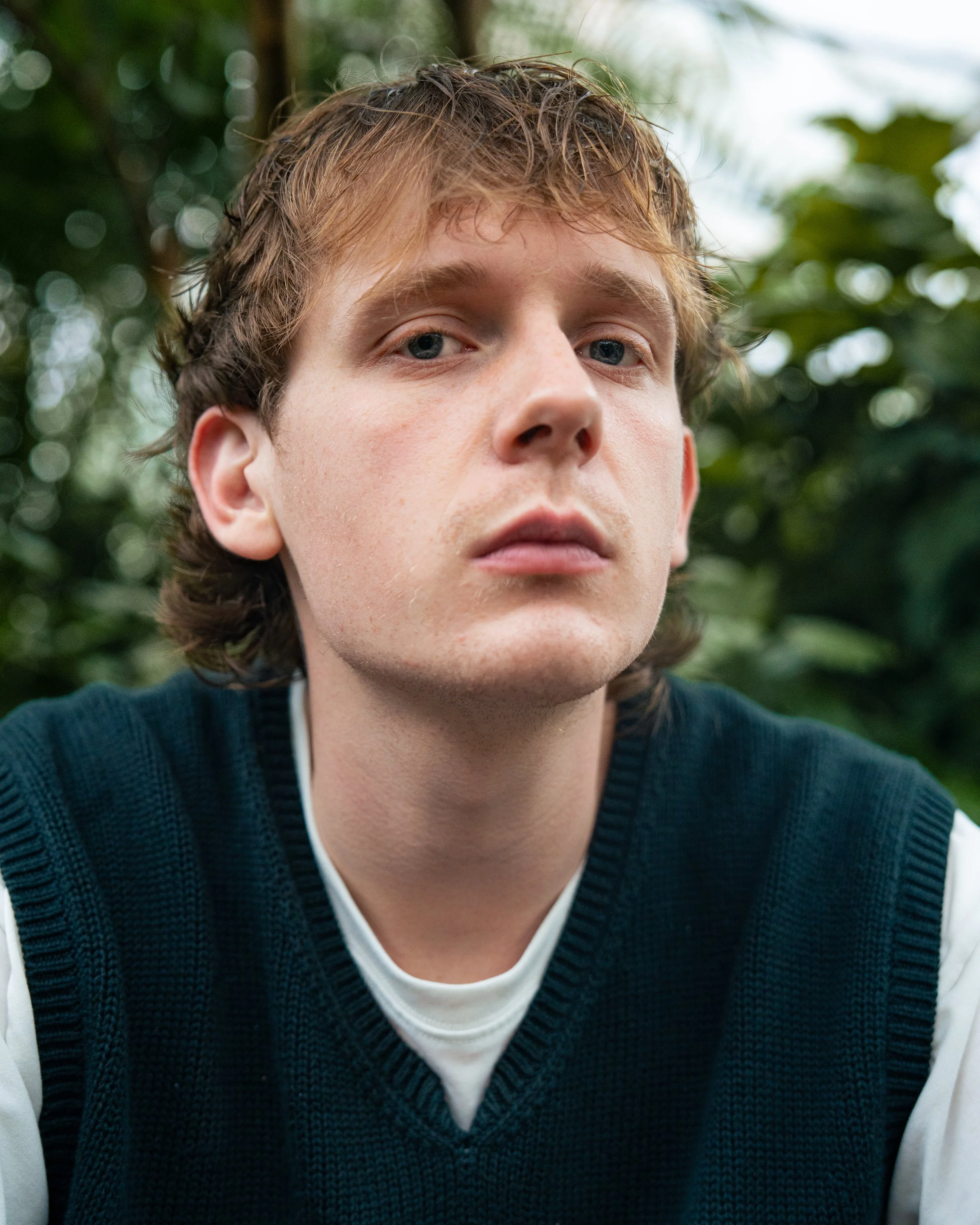 A young man with light skin, short wavy reddish-brown hair, and blue eyes, looking slightly upward with a neutral expression, outdoors with blurred green foliage in the background.