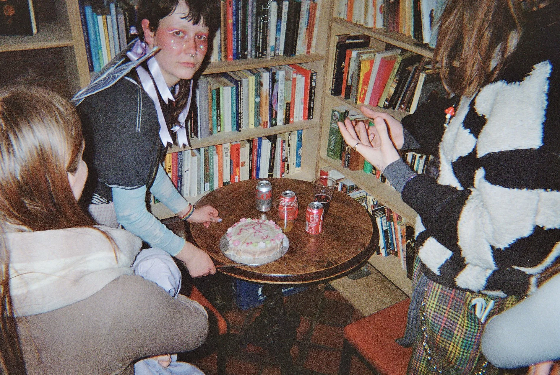 Group of young people gathered around a small round wooden table with a birthday cake and soda cans, in a room with bookshelves.