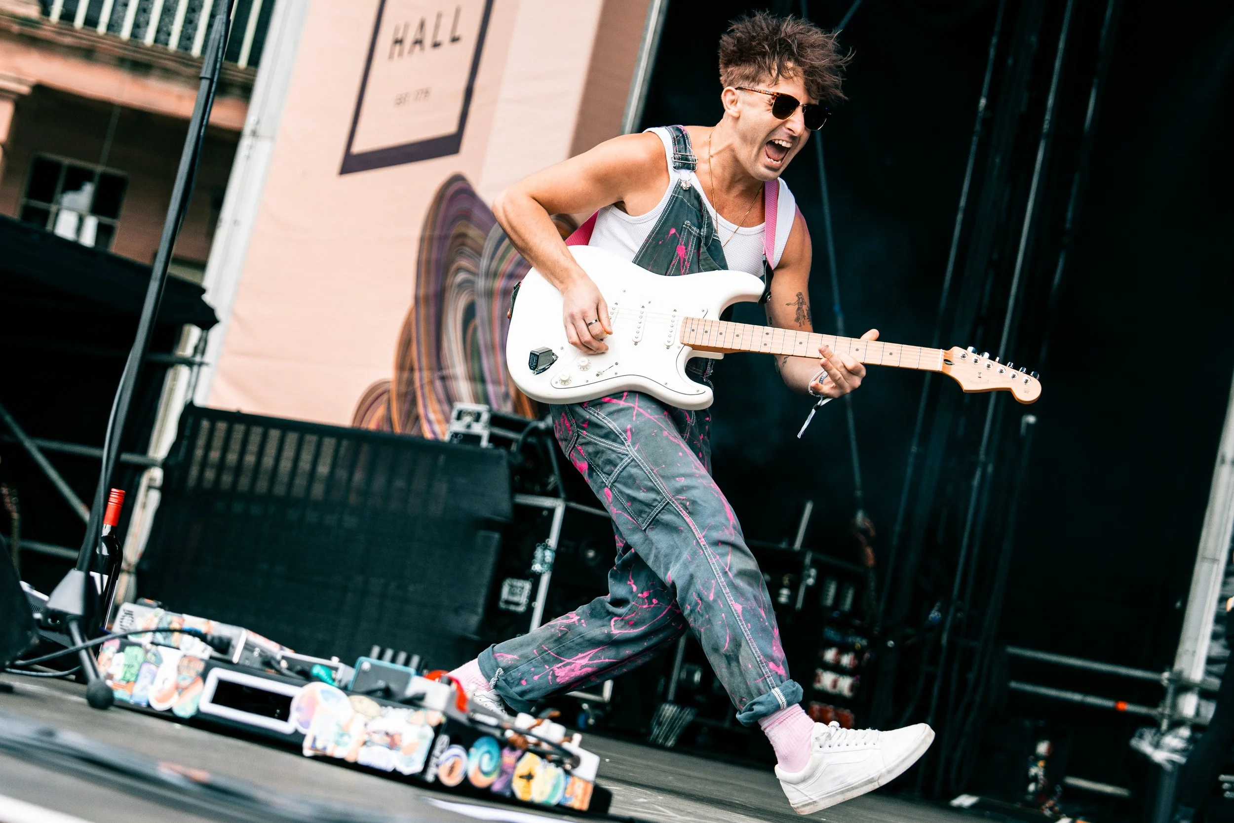 A person playing an electric guitar on stage during a live performance, wearing sunglasses, a sleeveless shirt, tie-dye pants, and white sneakers, with a backdrop that says 'HALL'.