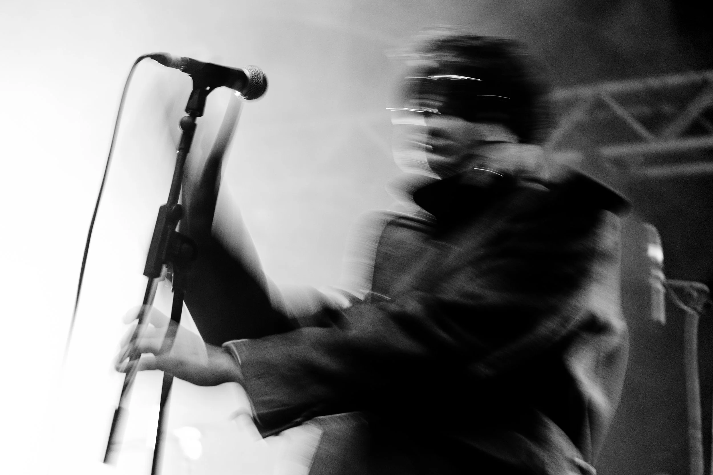 Blurred black and white photo of a man wearing sunglasses and a hoodie, holding a microphone on stage.