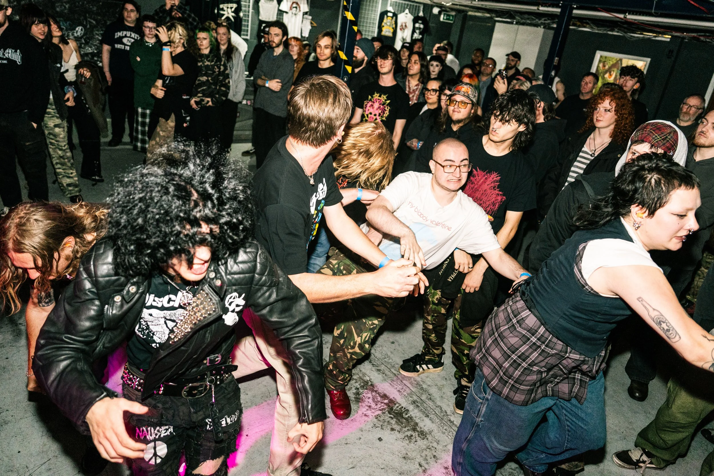 Individuals at a concert or music event are involved in a mosh pit, with some pushing and others dancing energetically, surrounded by a crowd watching the scene.