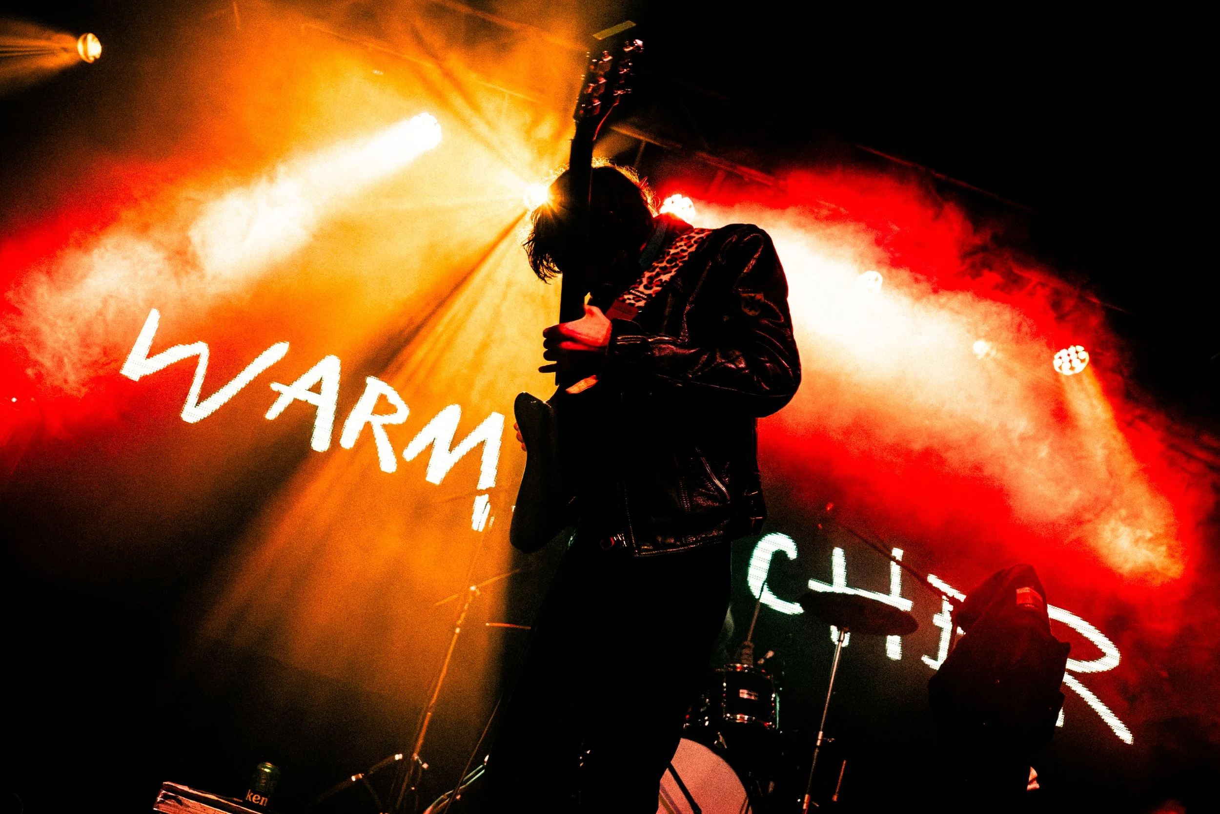 A musician playing an electric guitar on stage during a concert, backlit with orange and red stage lights, with smoky atmosphere and a drum set in the background.