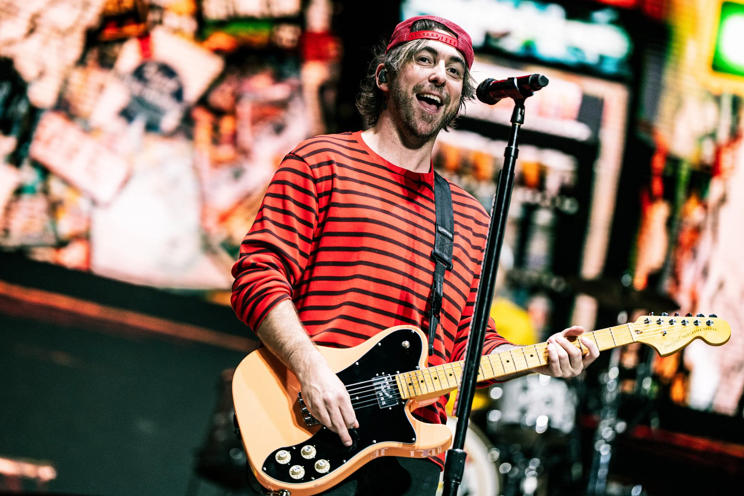 A man with long hair and a beard playing an electric guitar on stage, smiling, wearing a red and black striped shirt and a red baseball cap backward. The background is colorful and blurred.
