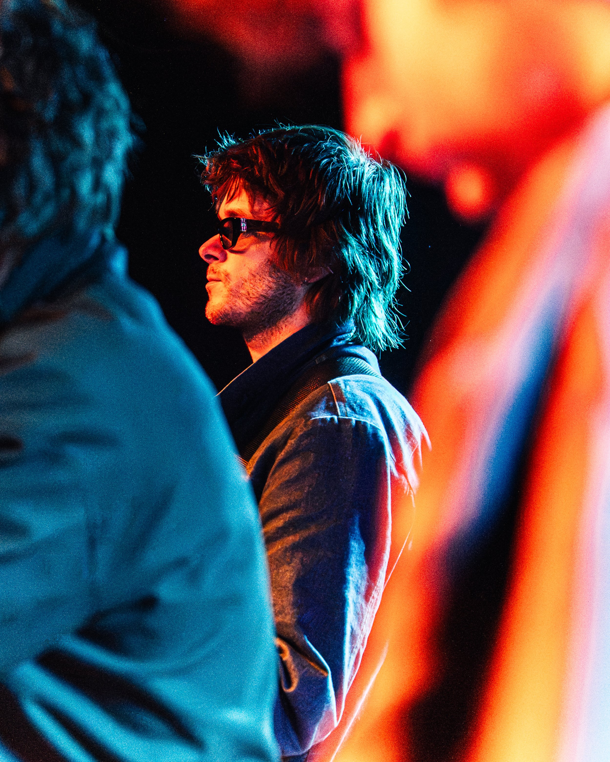 A side profile of a young man with medium-length hair, wearing glasses and a denim jacket, illuminated by colorful stage lighting in a dark setting.