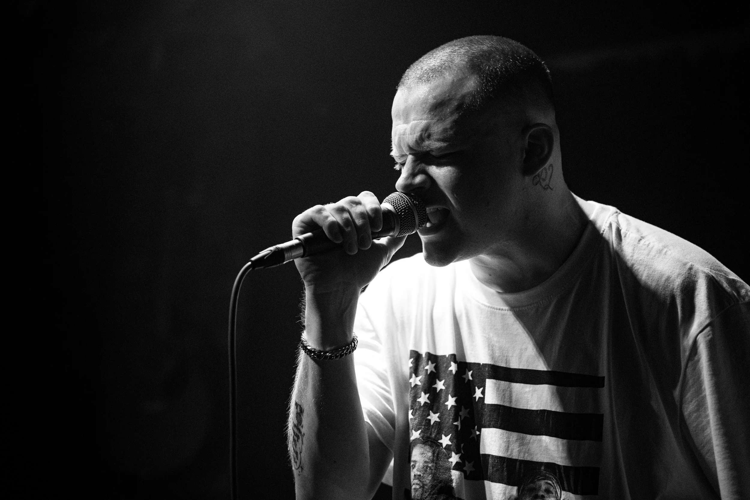 A black-and-white photo of a male singer passionately singing into a microphone, wearing a t-shirt with an American flag design, with tattoos and a bracelet visible.
