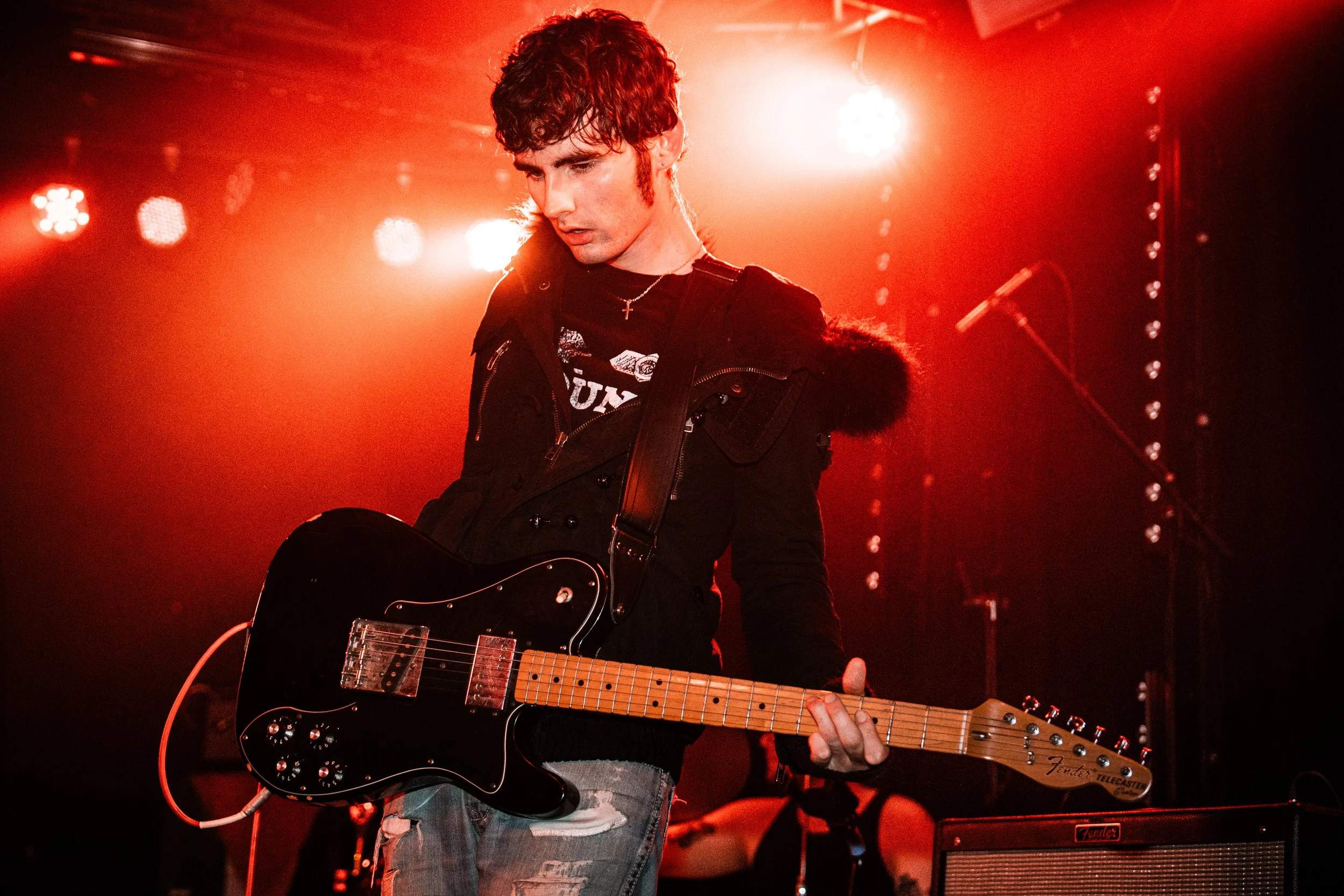 A young man playing an electric guitar on stage under red lighting, wearing a black jacket and distressed jeans.
