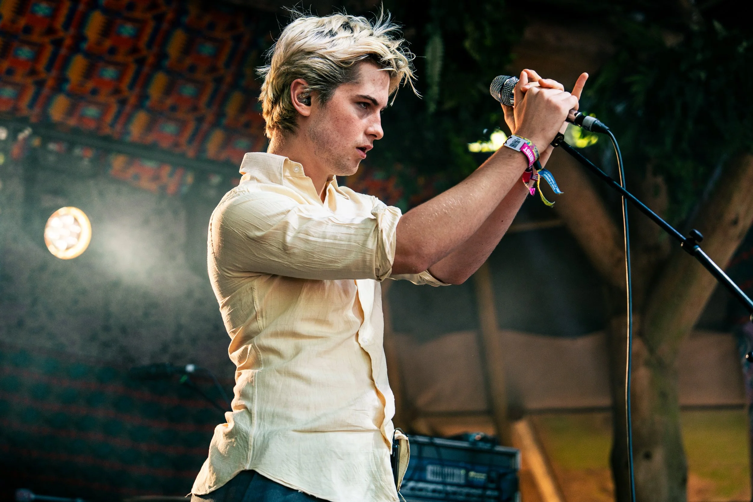 A young man with blonde hair, wearing a light yellow shirt with rolled-up sleeves, adjusting a microphone on a stand during a live performance or event. He is inside a wooden structure with colorful patterned ceiling and trees in the background.