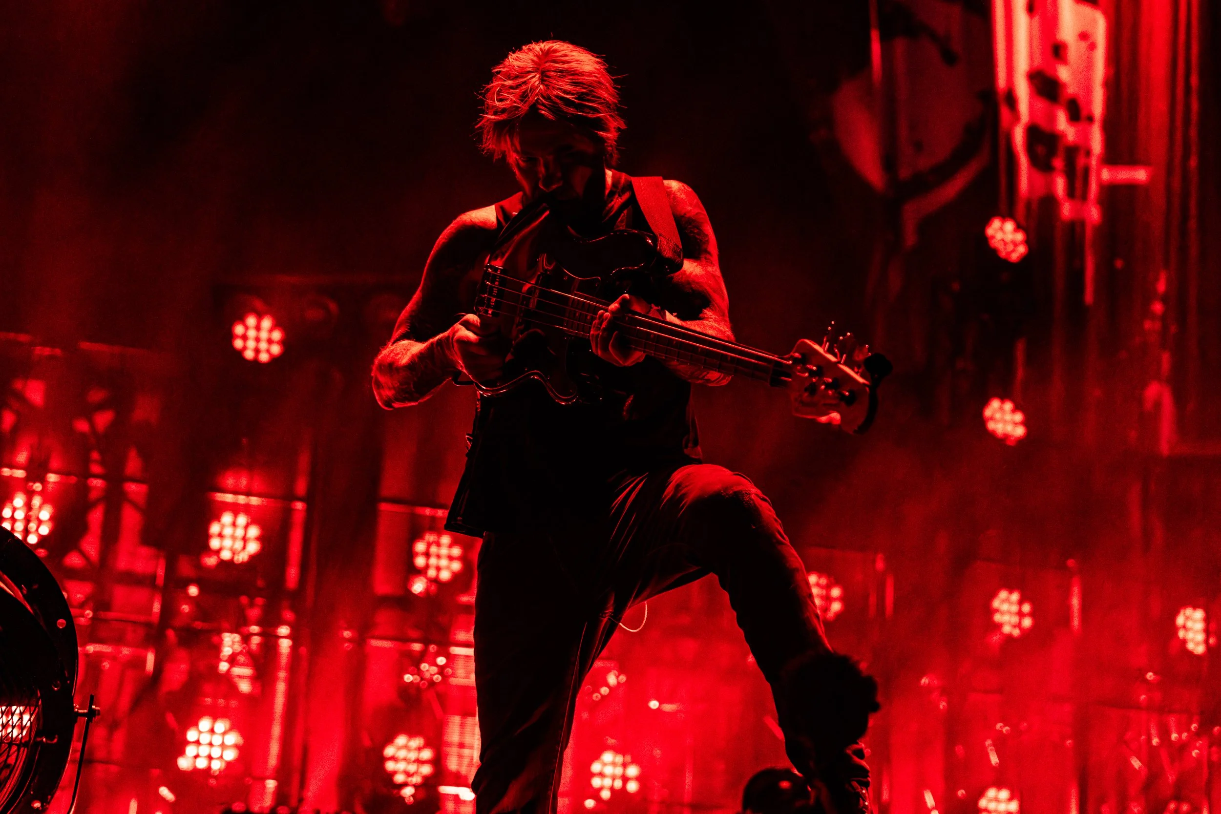 A musician playing an electric guitar on stage, illuminated by red stage lights, with a smoky background.