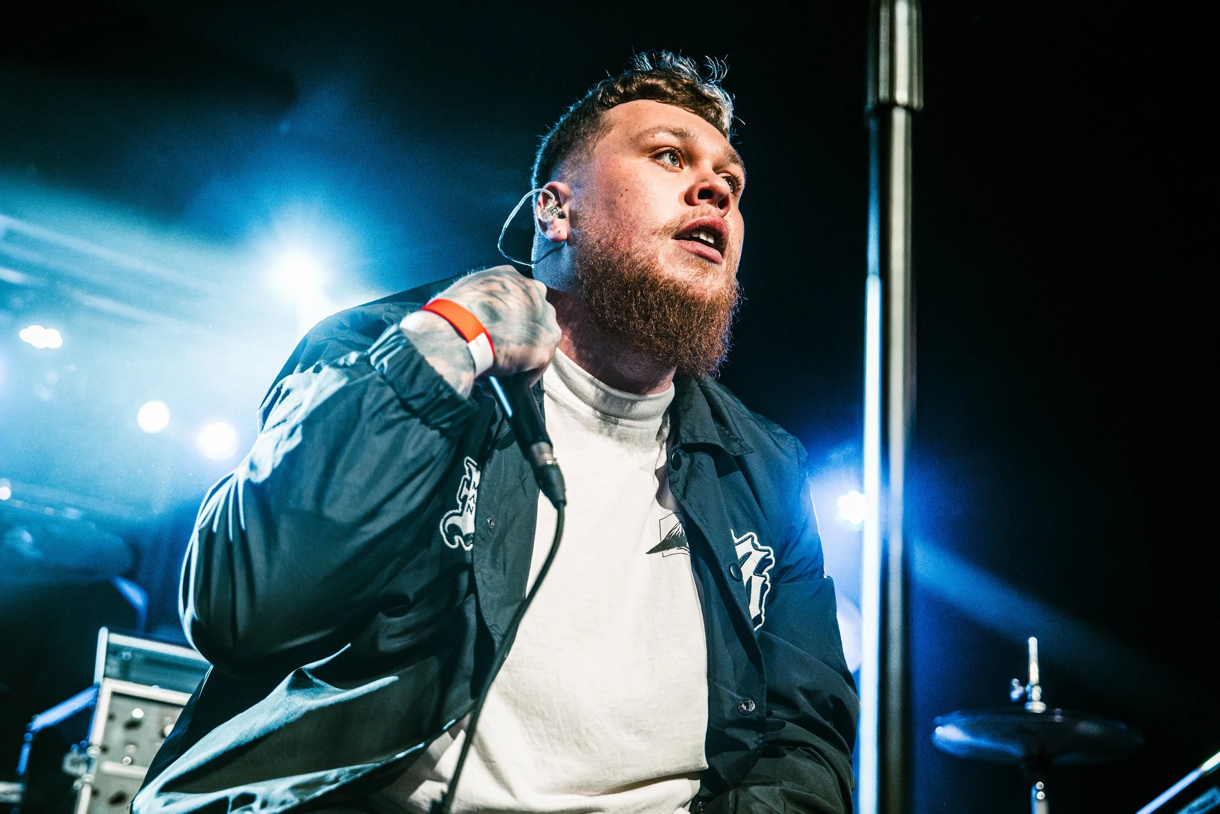 A male singer with a beard and tattoos singing into a microphone during a live performance on stage with blue and white stage lighting.