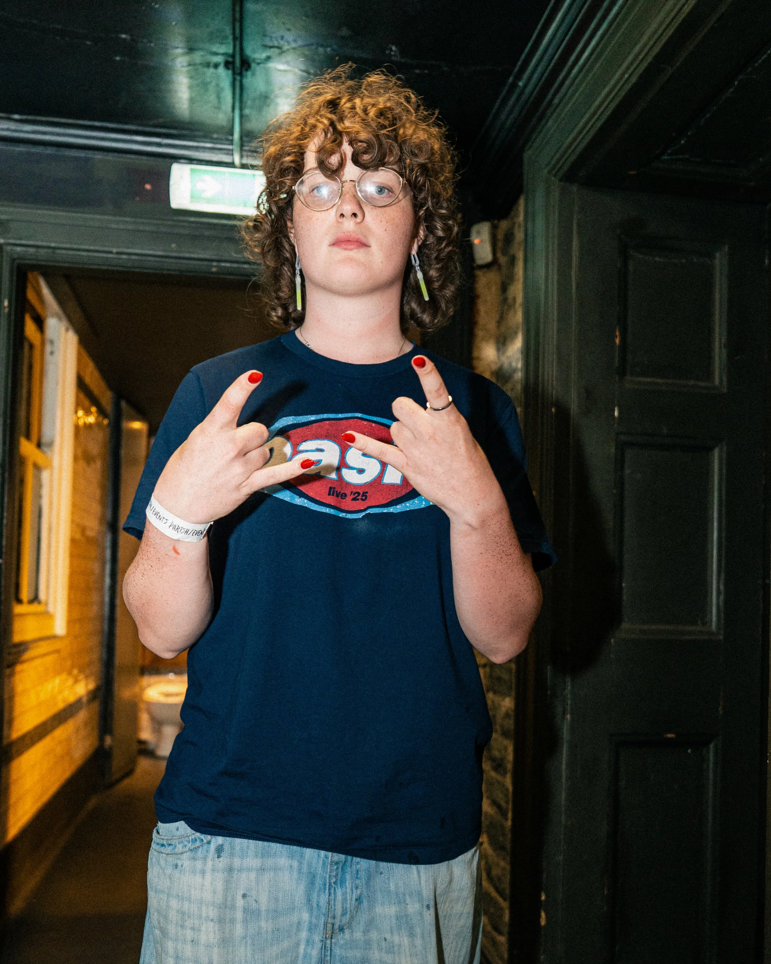 A young person with curly hair, glasses, and earrings, making a peace sign with both hands, wearing a blue T-shirt with a logo, standing indoors.