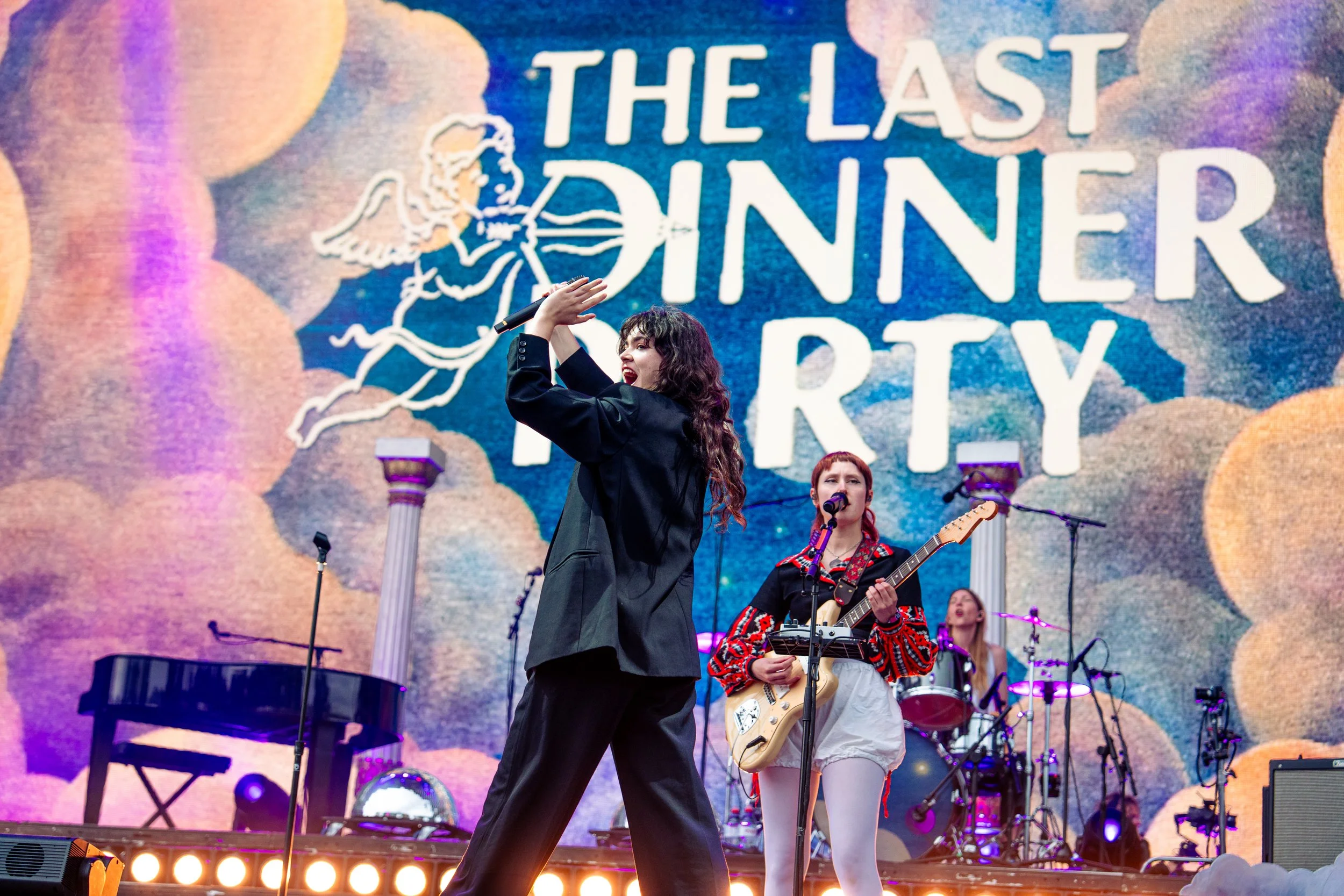 A band performs on stage during the last dinner party event, with a large animated background displaying clouds and the text 'The Last Dinner Party' and an angel figure.